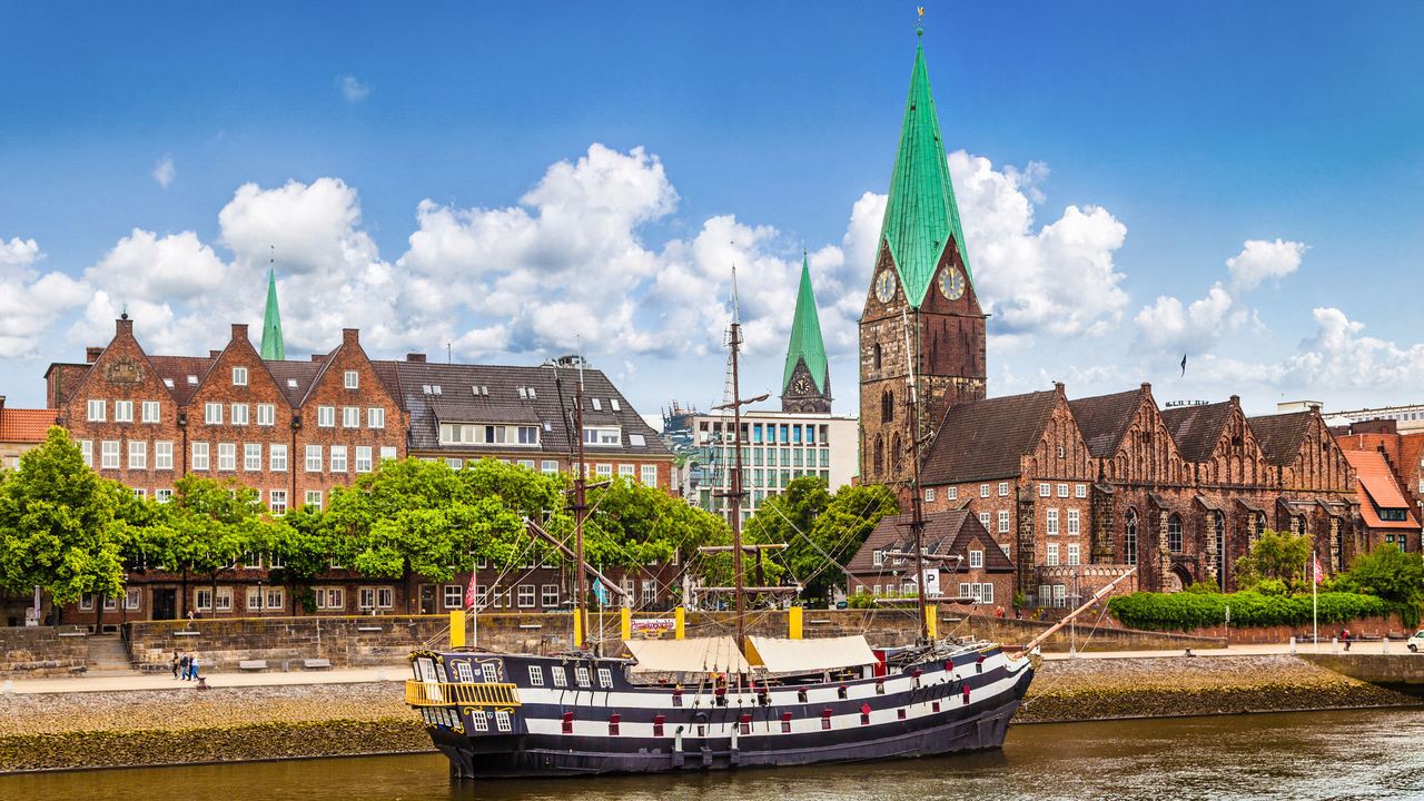 Blick auf eine historische Stadt mit roten Backsteingebäuden und hohen, grünen Kirchtürmen am Flussufer. Ein traditionelles Segelschiff liegt am Ufer, während in der Flussmitte ein Ruderboot mit vier Personen unterwegs ist. Der Himmel ist blau mit vereinzelten Wolken.