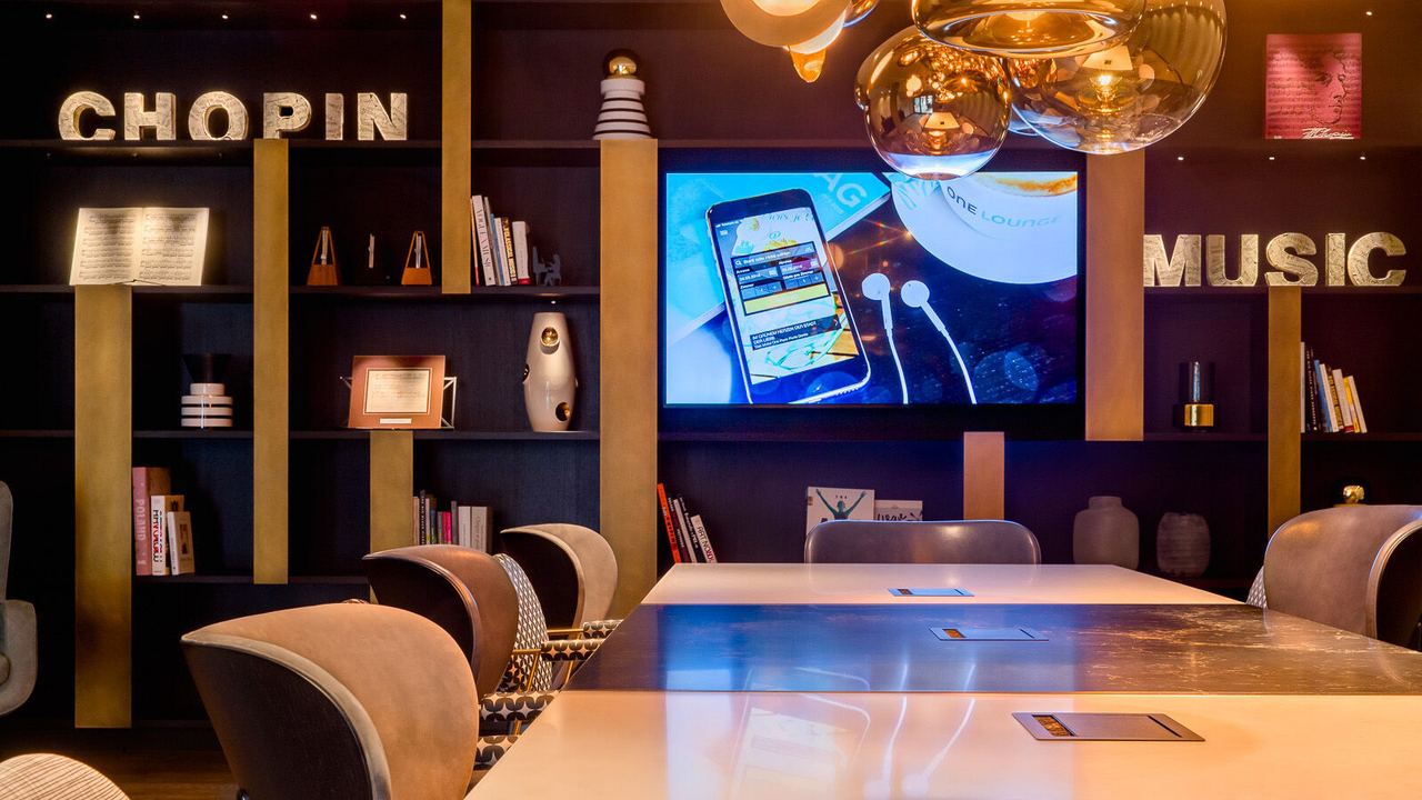 Motel One Besprechungsraum mit ergonomischen Stühlen, beleuchteten Regalen und dekorativen Schriftzügen „CHOPIN“ und „MUSIC“. Ein großer Bildschirm zeigt digitale Inhalte, das Ambiente wirkt kreativ und komfortabel.