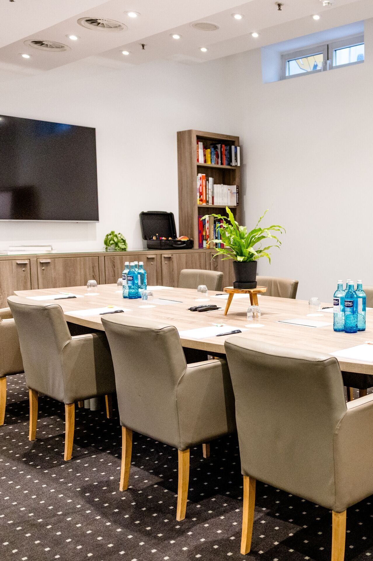 Hotel Frankfurt-Hauptbahnhof Motel One Conference room with large wooden table, upholstered armchairs, flipchart and television, ideal for meetings and presentations in a bright atmosphere.