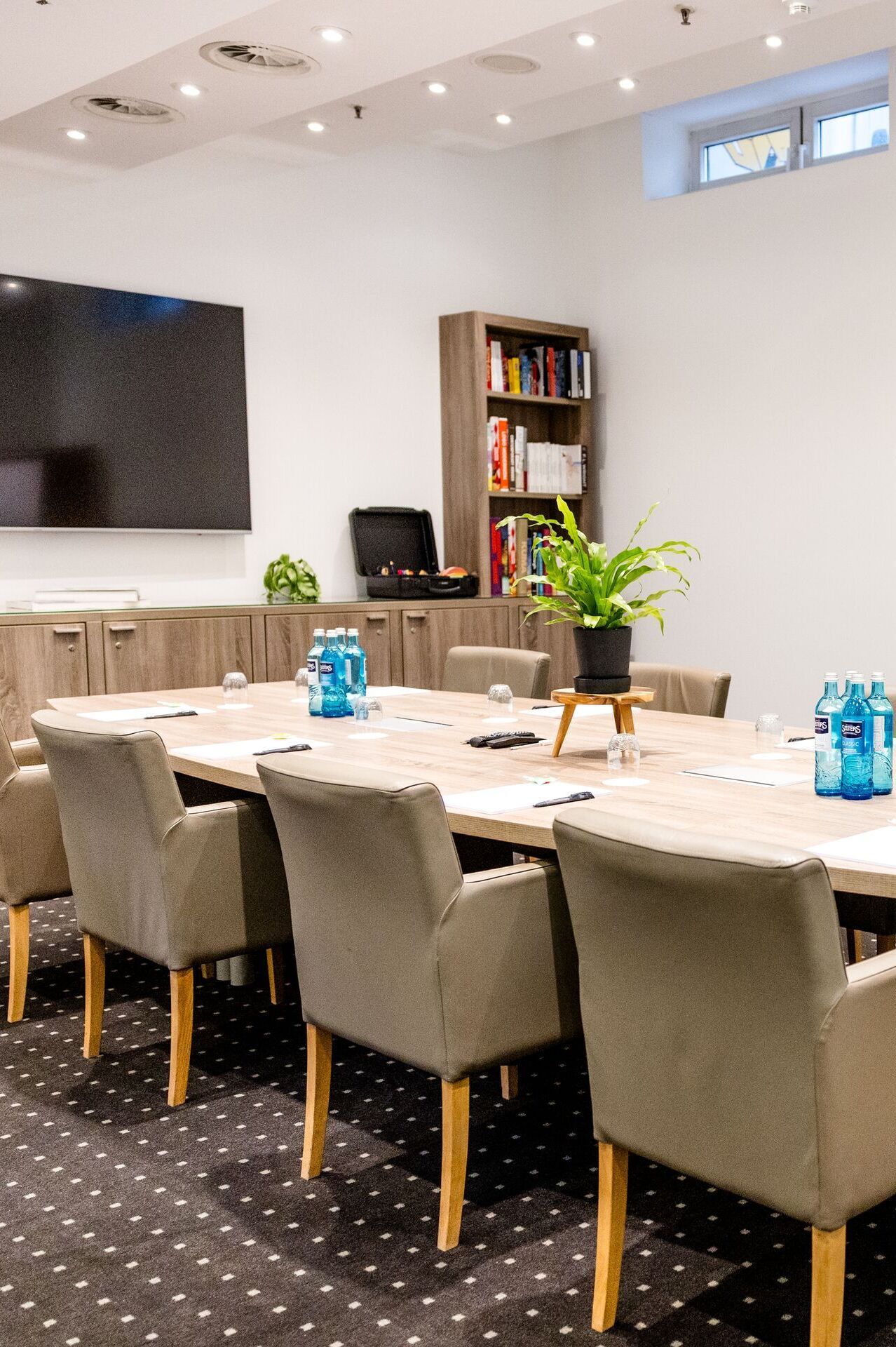 Hotel Frankfurt-Hauptbahnhof Motel One Conference room with large wooden table, upholstered armchairs, flipchart and television, ideal for meetings and presentations in a bright atmosphere.