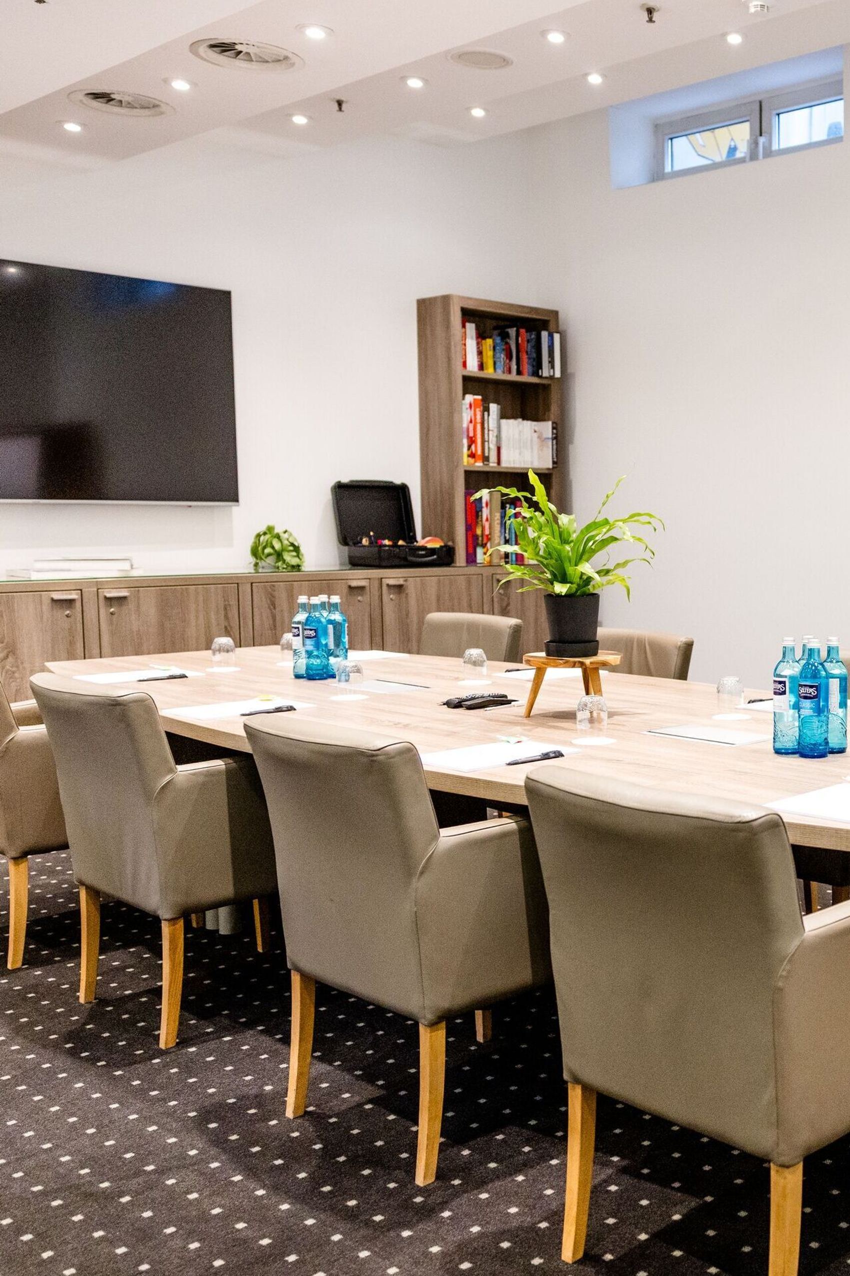 Hotel Frankfurt-Hauptbahnhof Motel One Conference room with large wooden table, upholstered armchairs, flipchart and television, ideal for meetings and presentations in a bright atmosphere.
