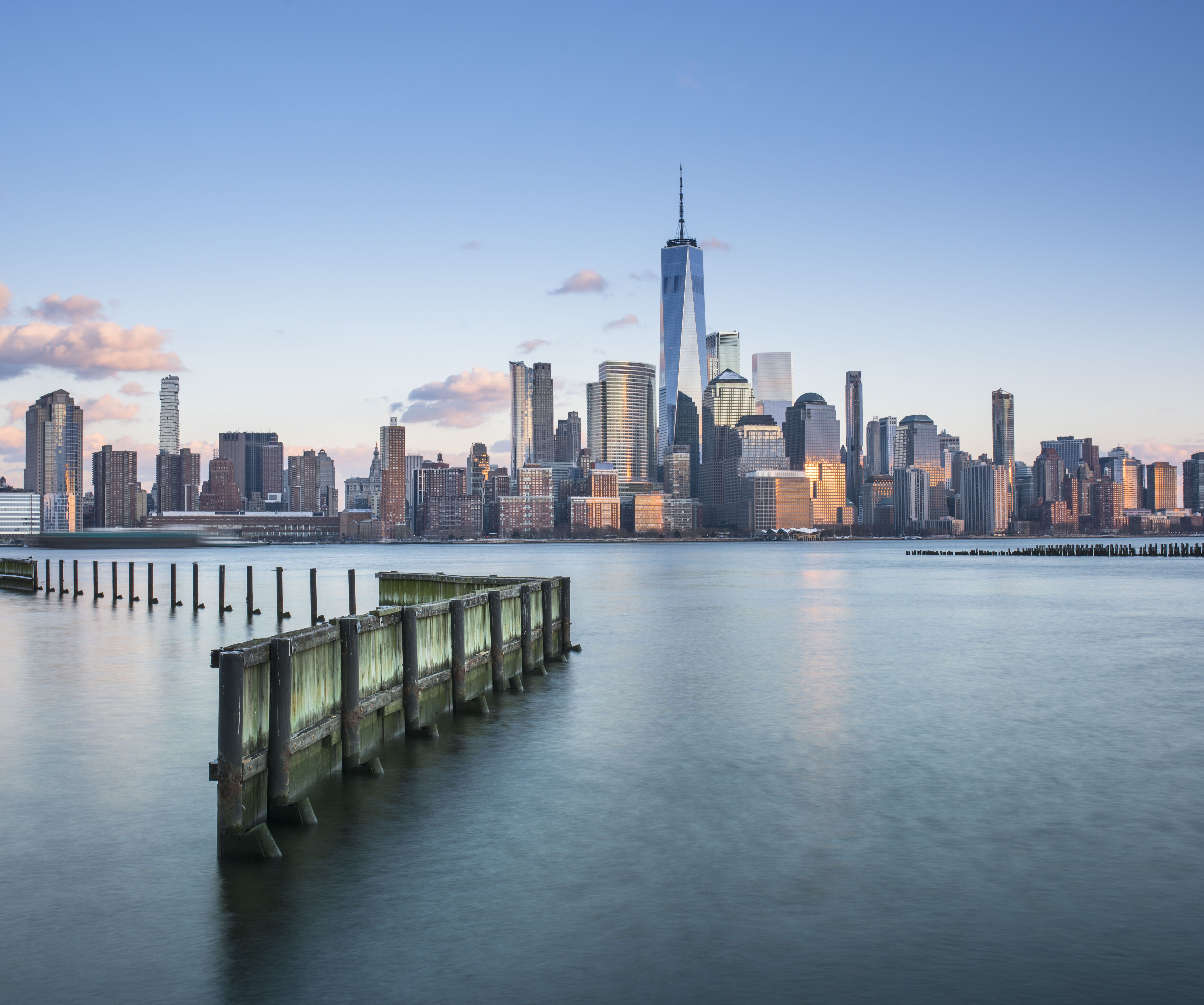 Blick auf die Skyline von Manhattan bei klarem Himmel, mit dem markanten One World Trade Center und ruhigem Wasser im Vordergrund, das eine alte Holzpier umspült.