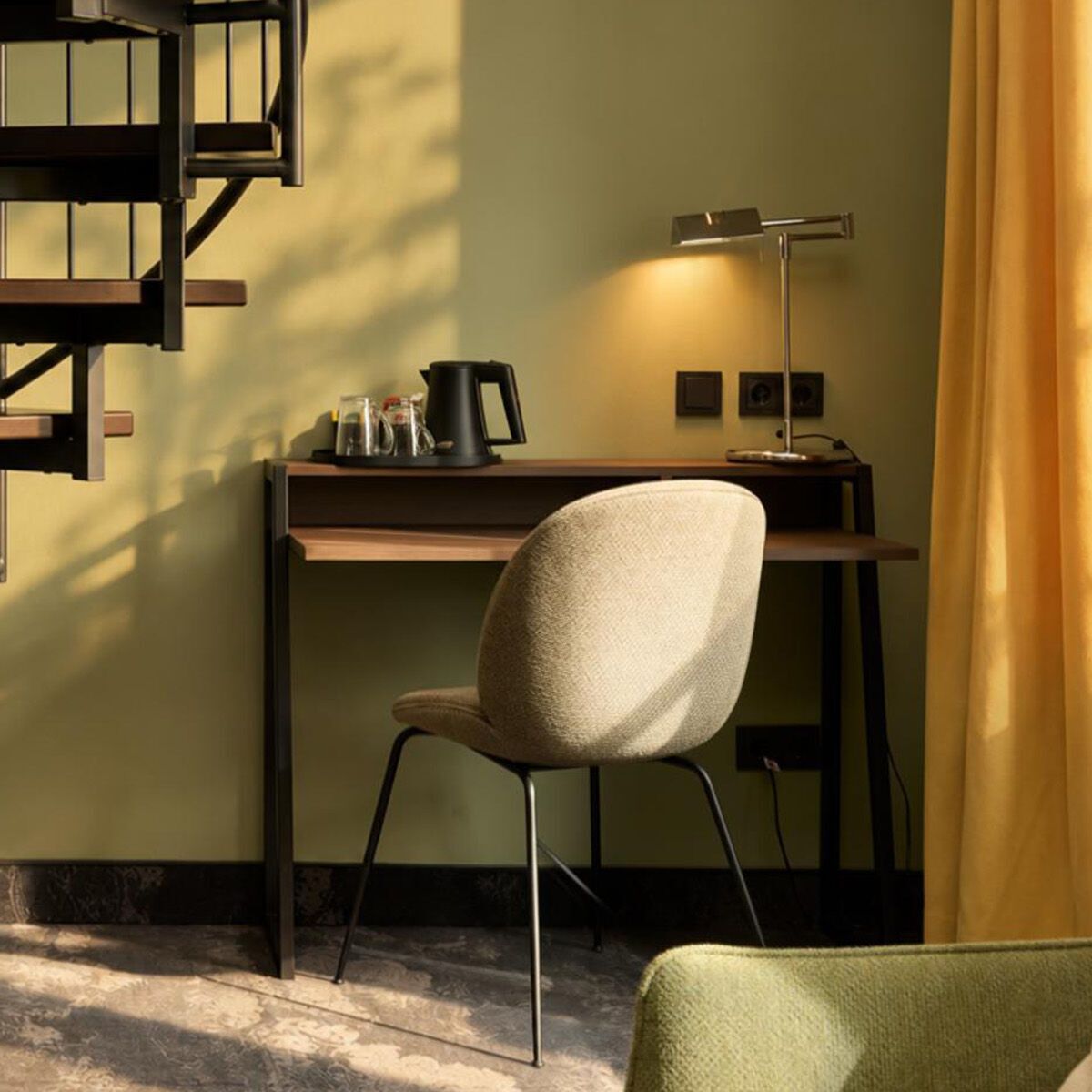 Modernes Zimmer mit gemütlichem Arbeitsplatz, Schreibtischlampe und Hydromaschine. Neben dem Schreibtisch führt eine schwarze Wendeltreppe nach oben. Sanftes Tageslicht fällt durch ein Fenster mit gelben Vorhängen.