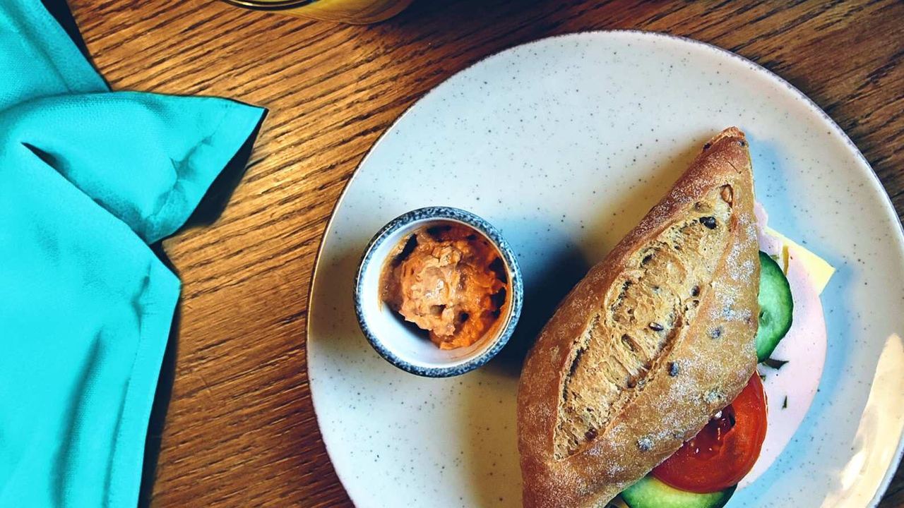 Frühstücksteller mit frischem Vollkornbrötchen, Tomaten- und Gurkenscheiben, einem kleinen Becher mit Aufstrich, dazu ein Glas Orangensaft und eine Tasse Kaffee auf Holztisch.