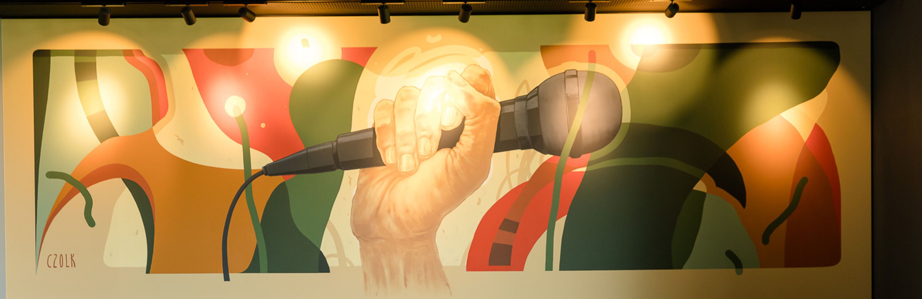 Hand hält ein Mikrofon vor einem farbenfrohen, abstrakten Wandbild mit warmem Licht