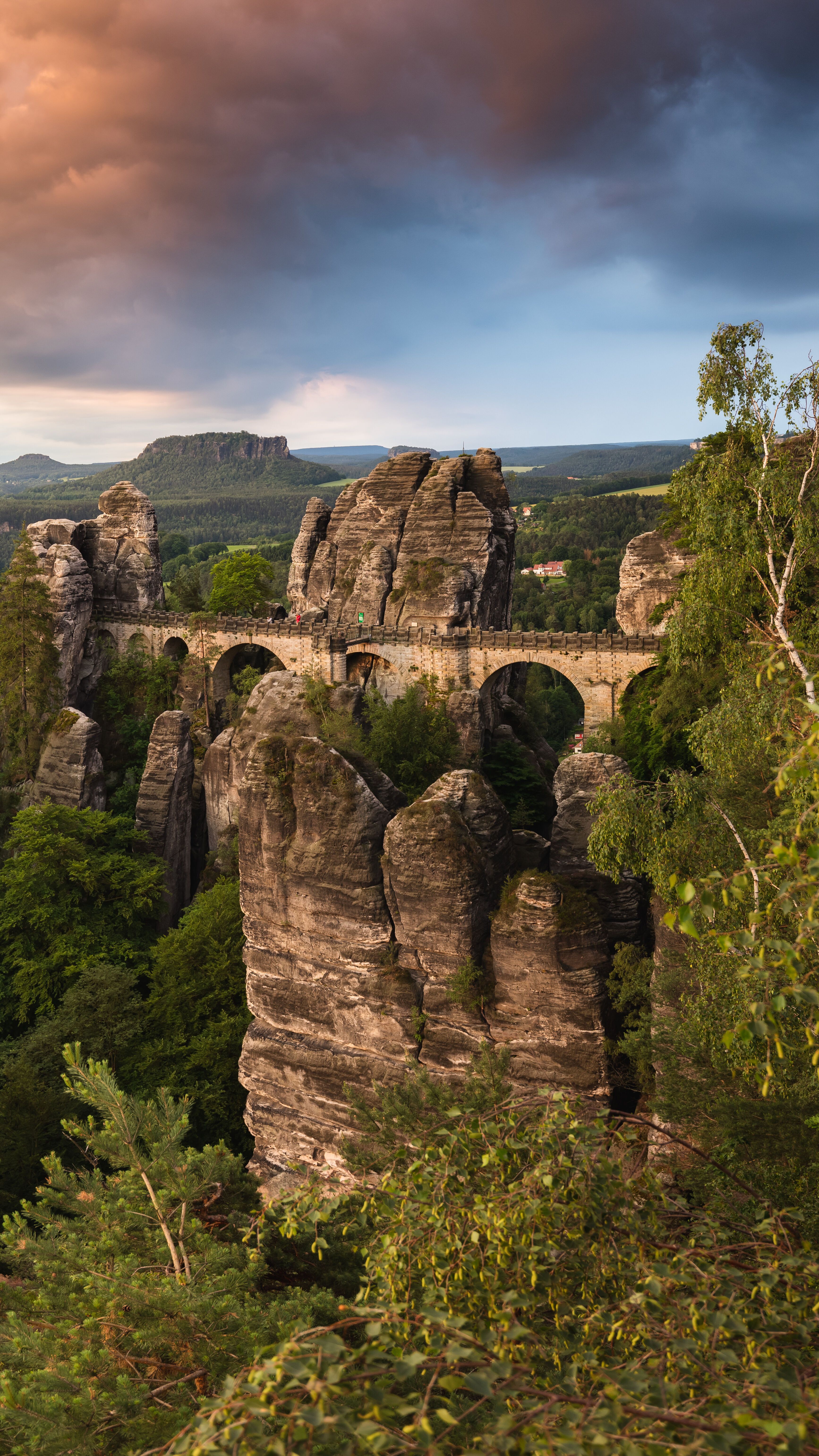 Felsformationen der Sächsischen Schweiz mit der Basteibrücke, umgeben von dichtem Wald, unter dramatischem Abendhimmel. Panoramablick auf die einzigartige Landschaft und natürliche Schönheit der Region.