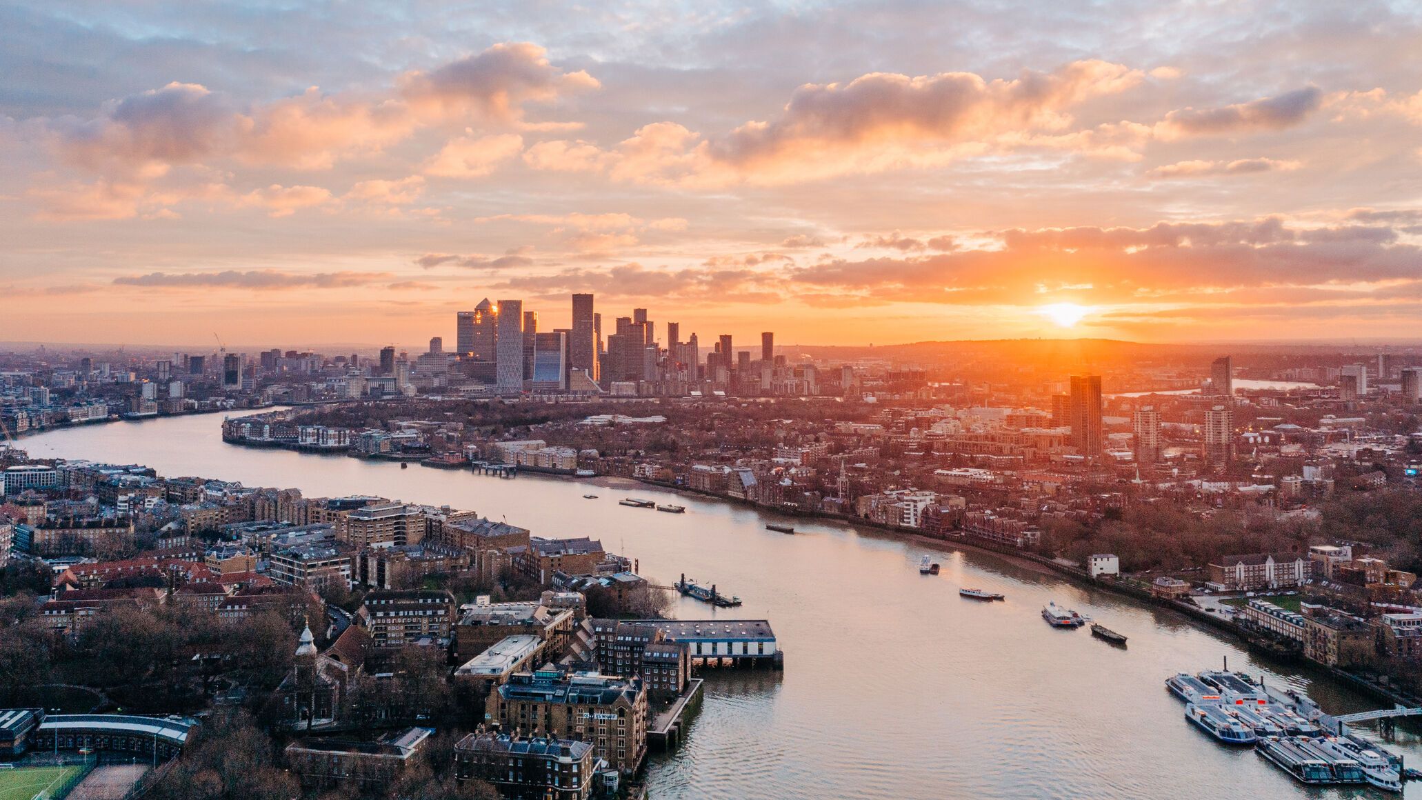 Sonnenuntergang über einer großen Stadt mit modernen Hochhäusern und einem breiten Fluss, der sich durch urbanes Gebiet mit Gebäuden und Booten schlängelt. Der Himmel zeigt warme Farben und vereint Stadtleben mit natürlicher Atmosphäre.