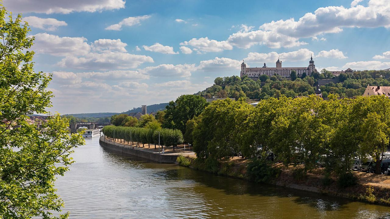 Blick auf einen ruhigen Fluss mit bewaldetem Ufer und einer historischen Burg auf einem Hügel unter blauem Himmel mit weißen Wolken.