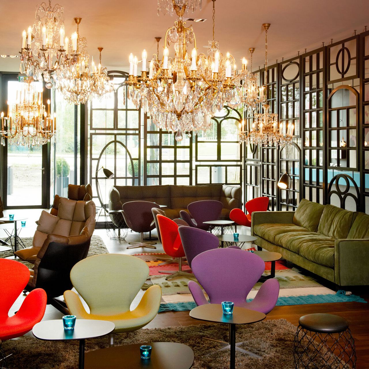 Heller, eleganter Motel One Lounge-Bereich mit Kronleuchtern, bunten Designerstühlen und gemütlichen Sofas, umgeben von großen Fenstern und modernen Raumteilern.