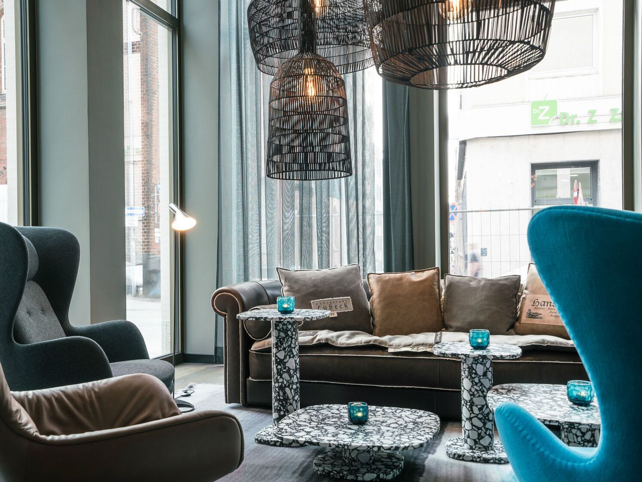 Gemütliche One Loungemöbel mit einem braunen Ledersofa, Sesseln in Grau und Blau, schwarzen Hängeleuchten und kleinen Tischen.