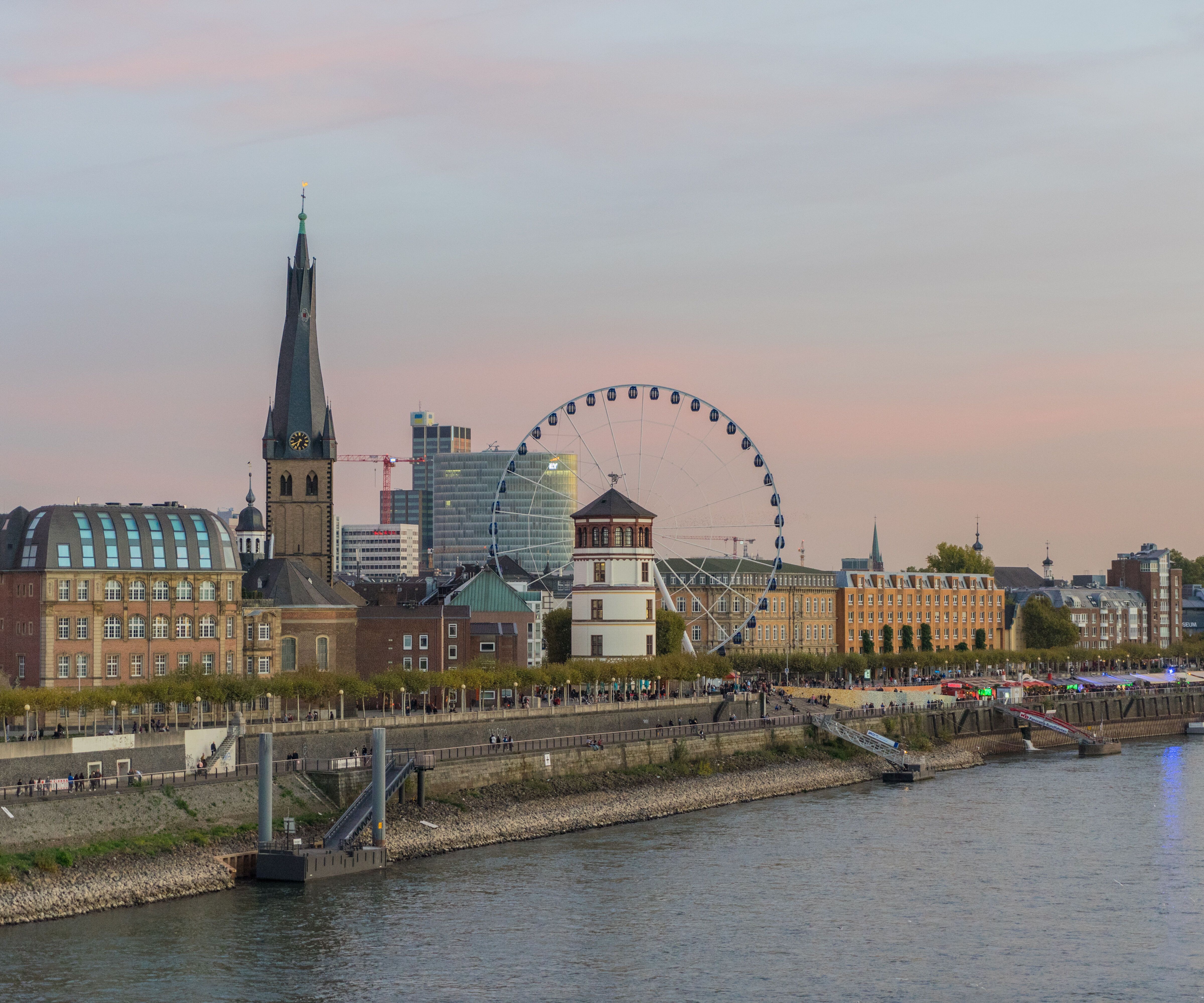 Blick auf eine Flussuferpromenade bei Sonnenuntergang mit historischer Stadtkulisse, einem Riesenrad und einem markanten Kirchturm, die eine lebendige und einladende Atmosphäre vermitteln.