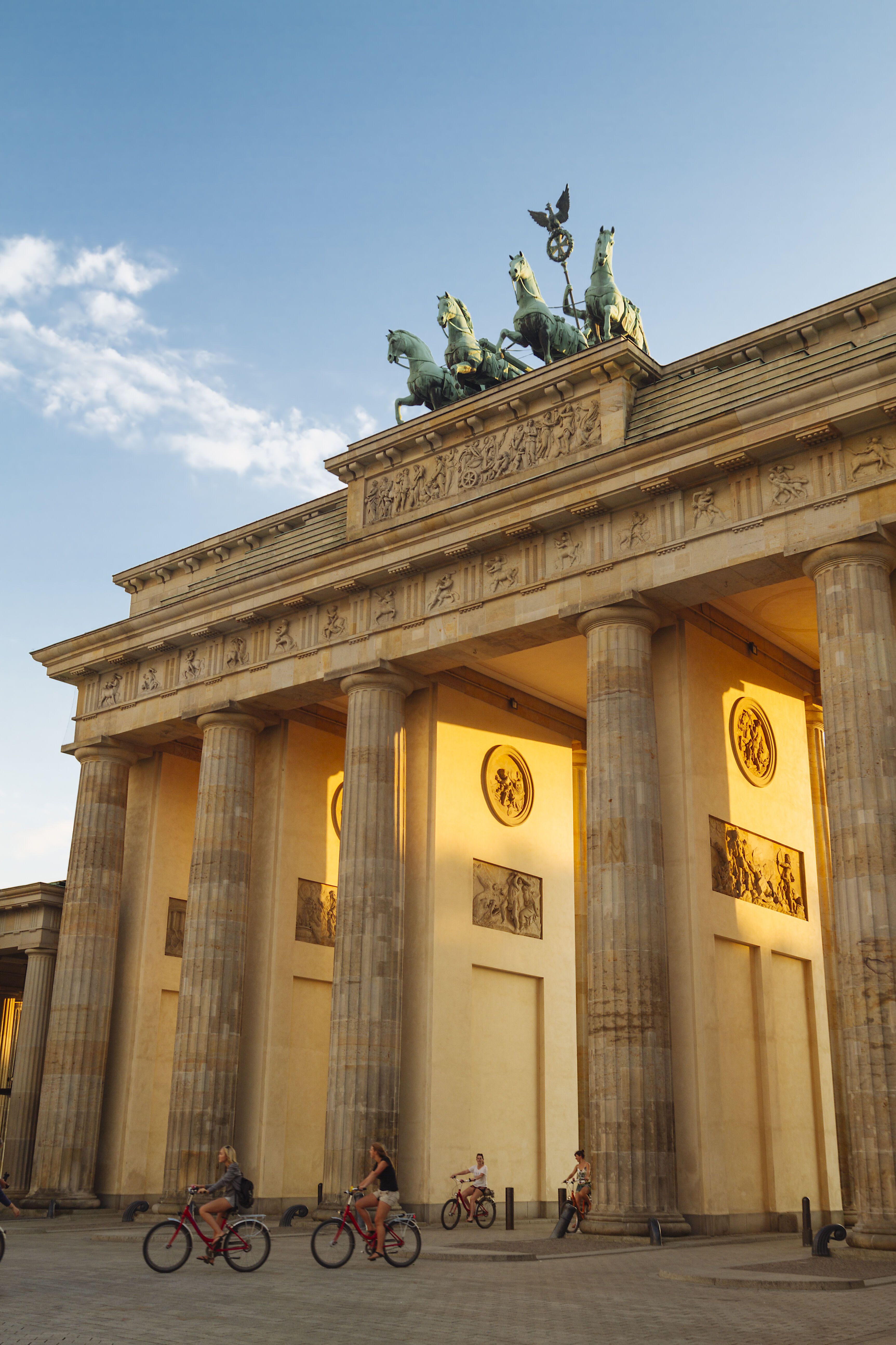 Brandenburger Tor in Berlin bei Sonnenuntergang, beleuchtet, mit mehreren Radfahrern vor den hohen Säulen und der Quadriga auf dem Dach.