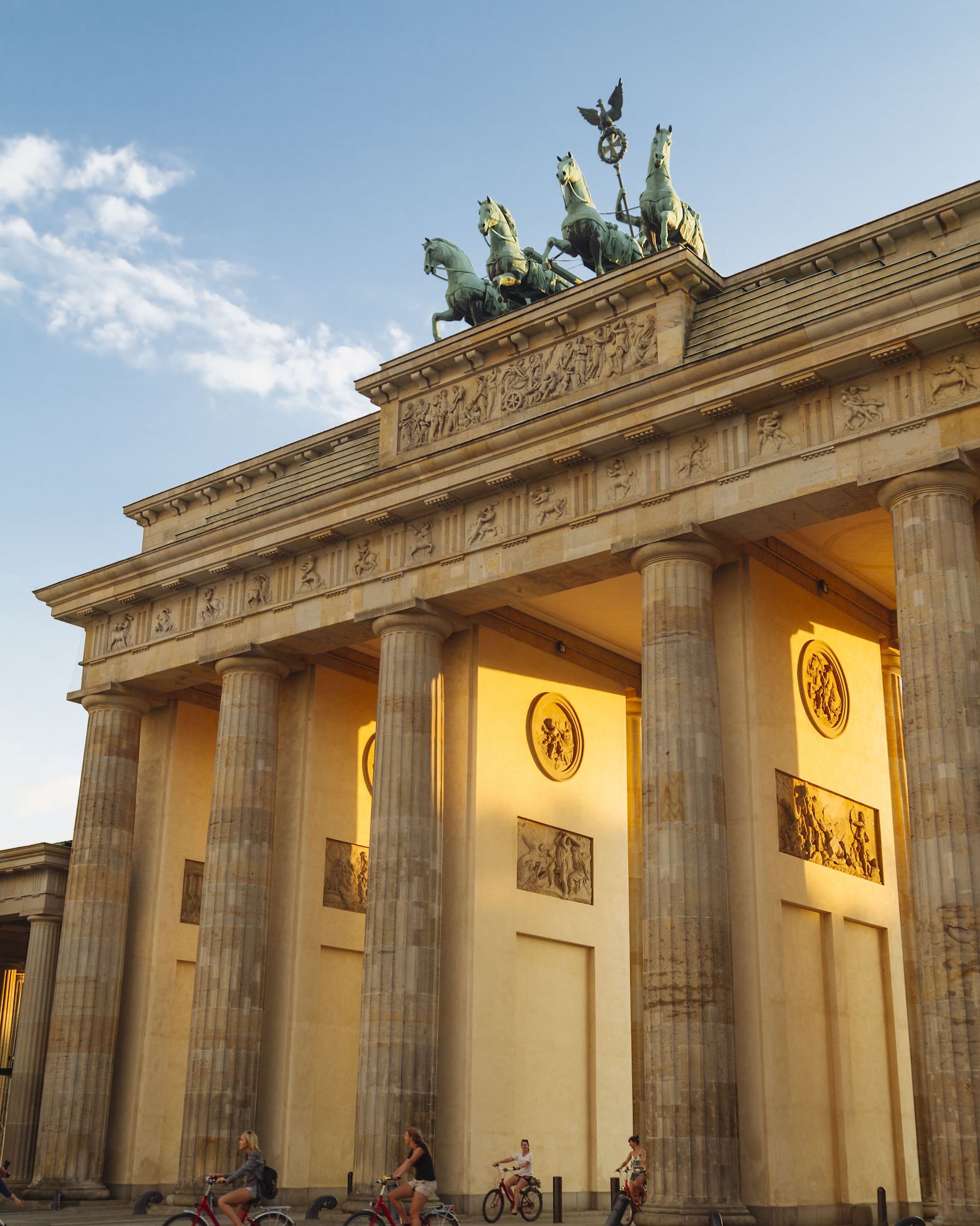Brandenburger Tor in Berlin bei Sonnenuntergang, beleuchtet, mit mehreren Radfahrern vor den hohen Säulen und der Quadriga auf dem Dach.