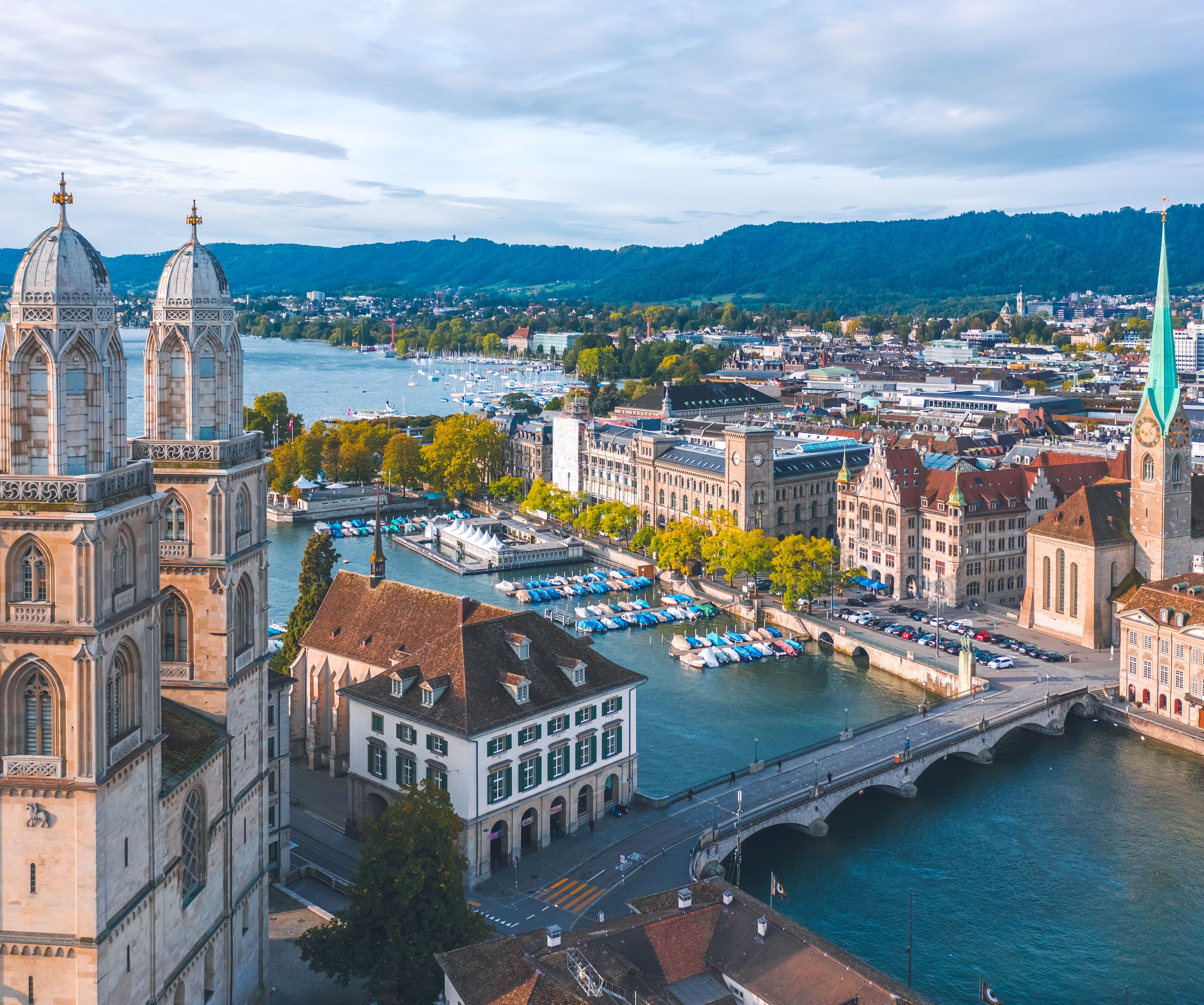 Blick auf Zürich mit der Limmat, historischen Gebäuden, den Zwillingstürmen der Grossmünsterkirche und der steilen Turmspitze der Fraumünsterkirche, umgeben von Bergen und blauem Himmel.