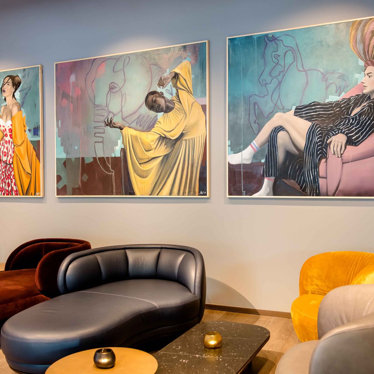 Stilvoll eingerichteter Lounge-Bereich mit farblich kontrastierenden Sitzmöbeln und drei ausdrucksstarken, zeitgenössischen Gemälden an der Wand.
