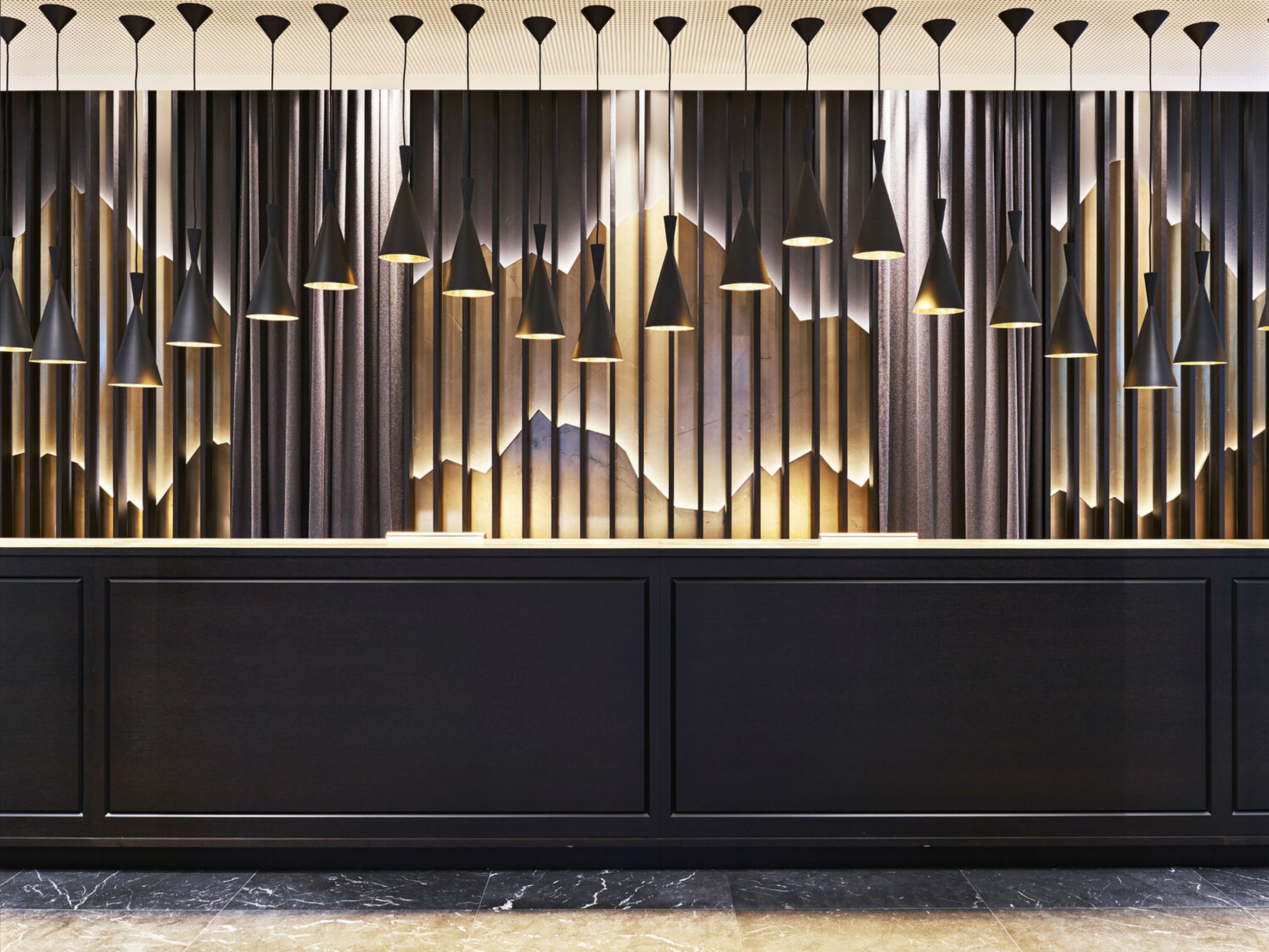 Motel One Hotelrezeption mit schwarzer Theke, darüber viele schlanke, schwarze Hängeleuchten in unterschiedlichen Längen. Elegante Wandgestaltung mit wellenförmigen, beleuchteten Strukturen hinter dunklen Vorhängen erzeugt ein stilvolles Ambiente.