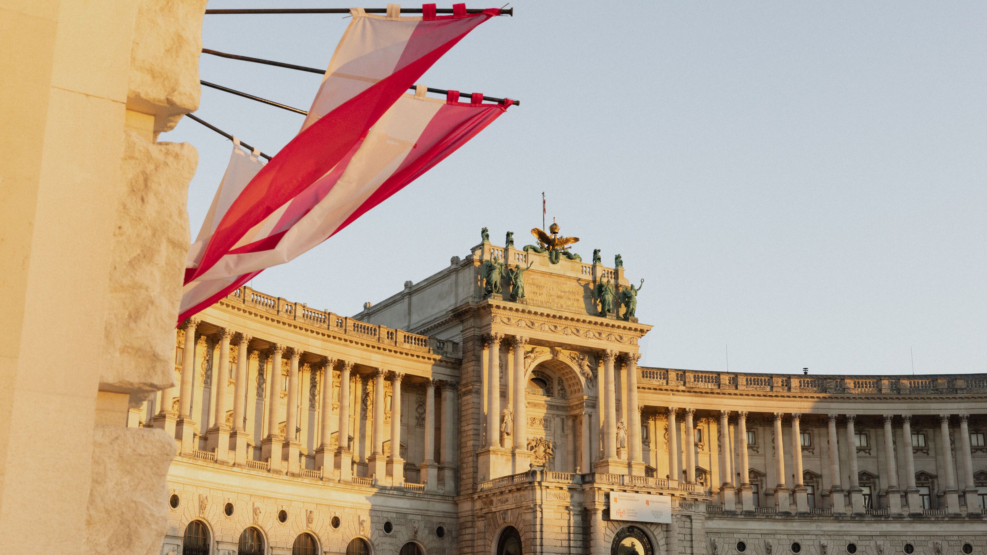 Historisches, prachtvolles Gebäude mit Säulen und Statuen bei warmem Abendlicht, davor hängende rot-weiß-rote Fahnen, die eine festliche Atmosphäre vermitteln.