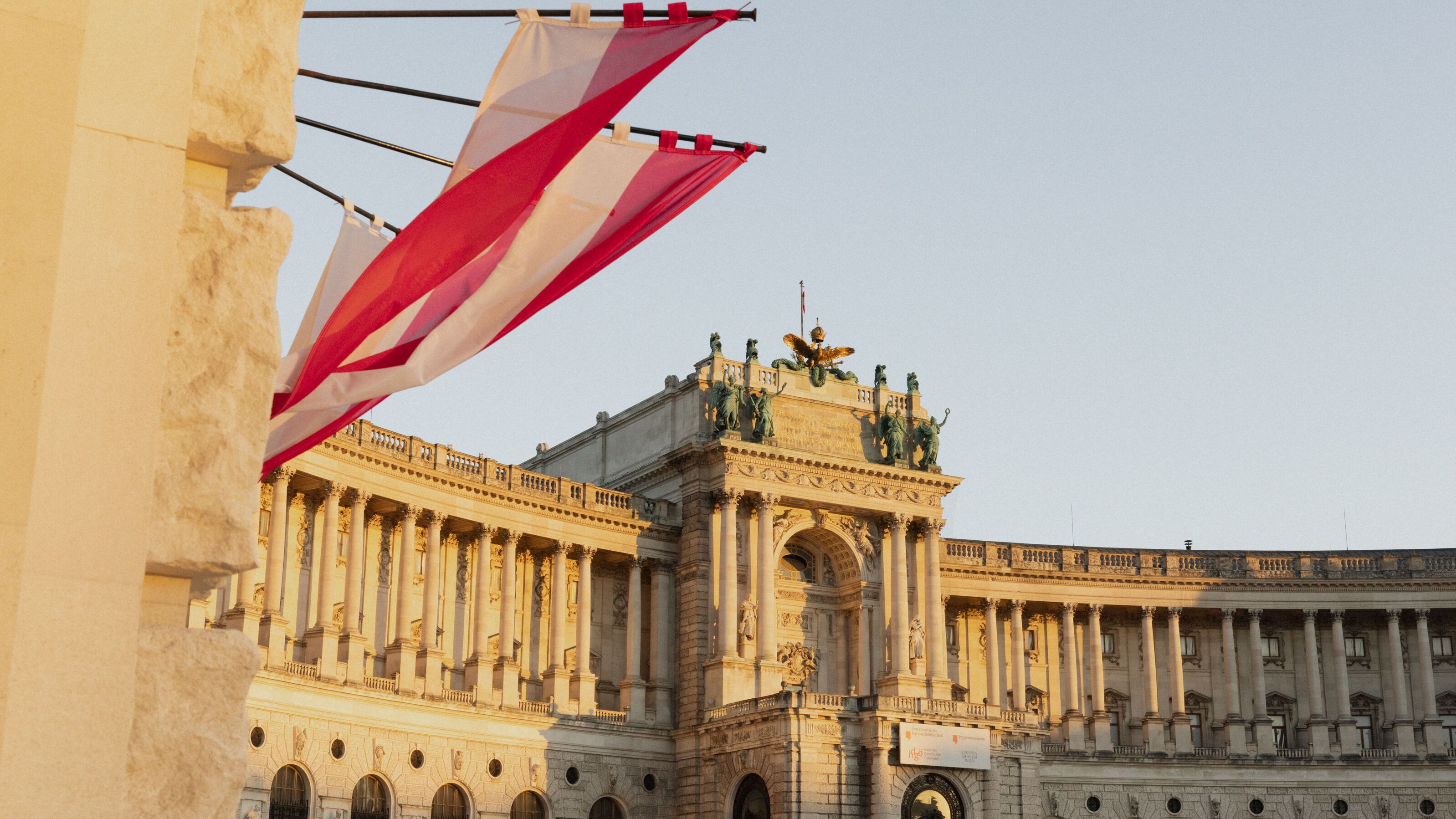 Historisches, prachtvolles Gebäude mit Säulen und Statuen bei warmem Abendlicht, davor hängende rot-weiß-rote Fahnen, die eine festliche Atmosphäre vermitteln.