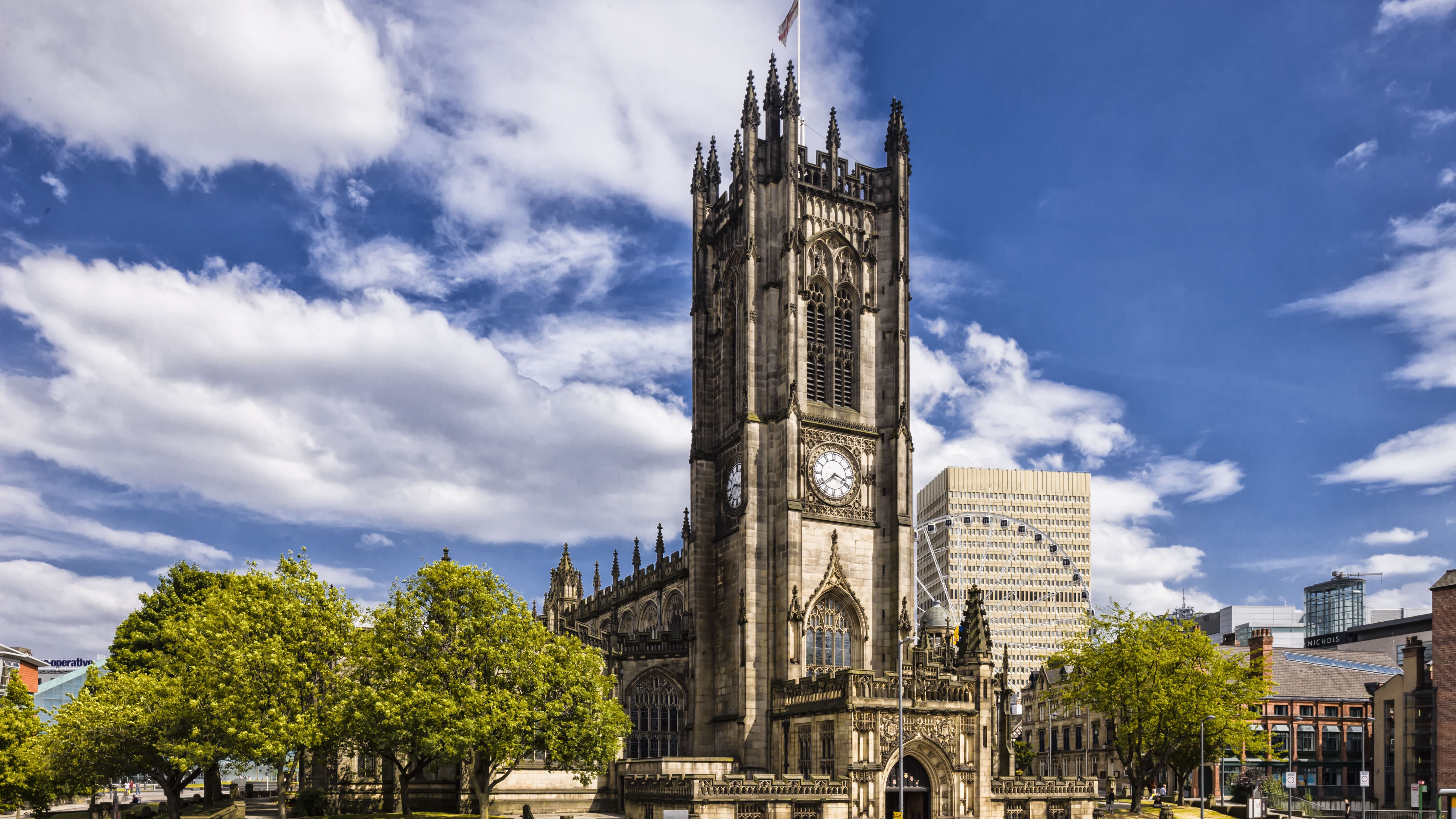 Gotisch inspirierte Kirche mit hohem Turm und Uhr, umgeben von Bäumen und städtischer Bebauung unter wechselnd bewölktem Himmel.