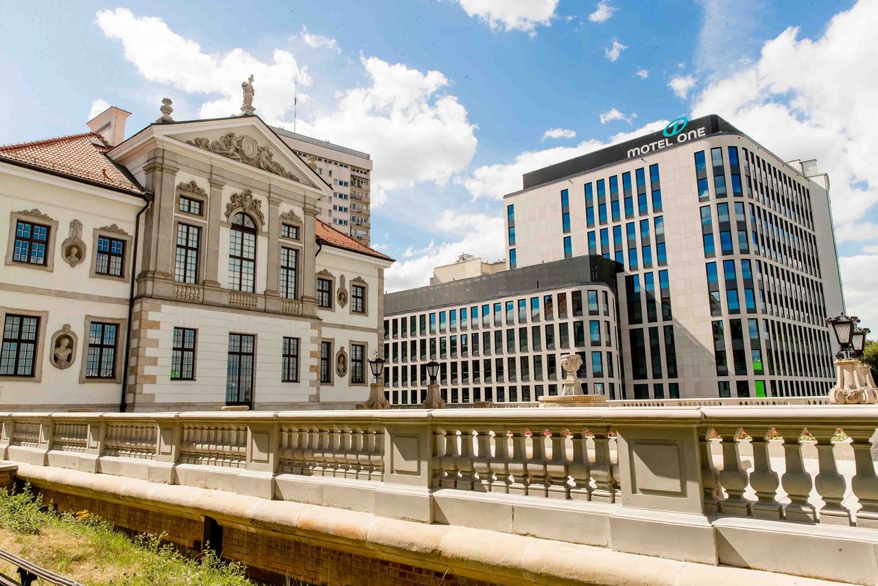 Historisches Gebäude und Motel One Hotel nebeneinander, strahlender Himmel, urbaner Stadteindruck, Kombination aus Tradition und zeitgenössischer Architektur.