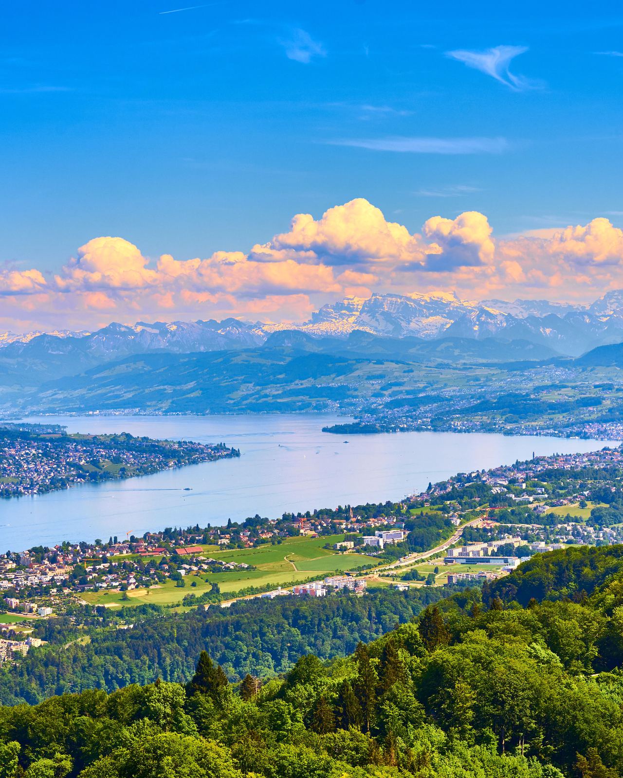 Weitläufige Panoramaansicht auf einen großen See, umgeben von grünen Hügeln und Städten, im Hintergrund majestätische Berge unter blauem Himmel mit weißen Wolken.