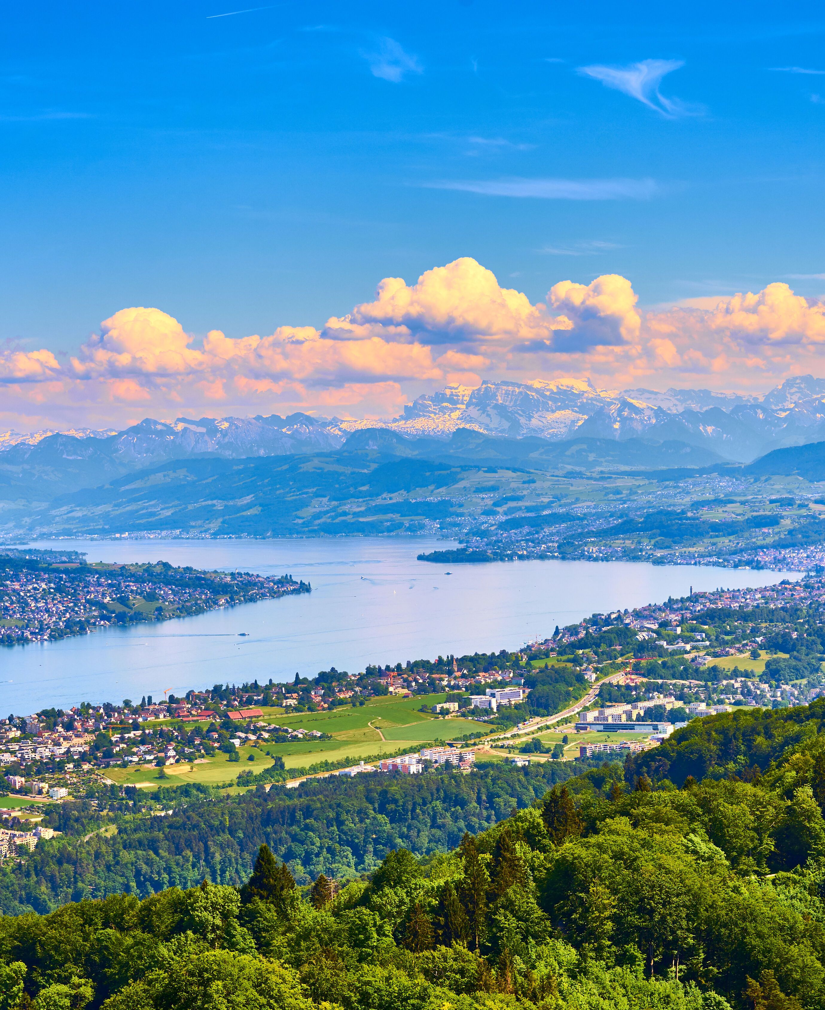 Weitläufige Panoramaansicht auf einen großen See, umgeben von grünen Hügeln und Städten, im Hintergrund majestätische Berge unter blauem Himmel mit weißen Wolken.