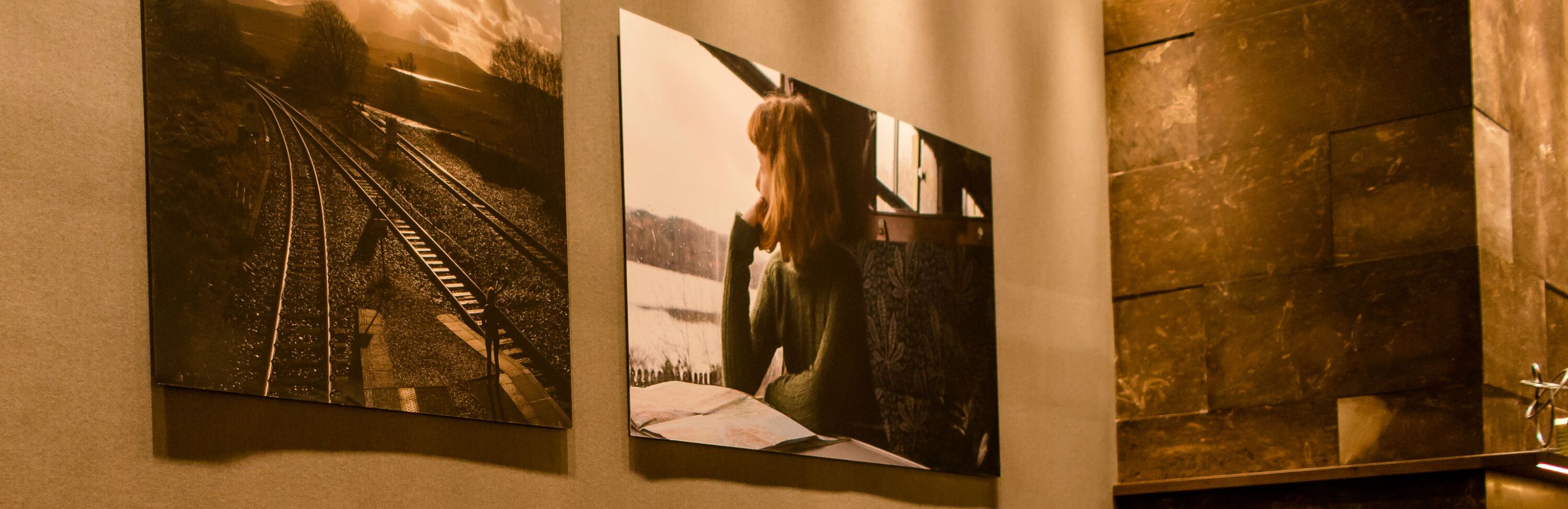 Zwei gerahmte Fotos hängen an einer Wand: eines zeigt Eisenbahnschienen im Sonnenuntergang, das andere eine nachdenkliche Frau an einem Fenster mit Blick auf einen See.