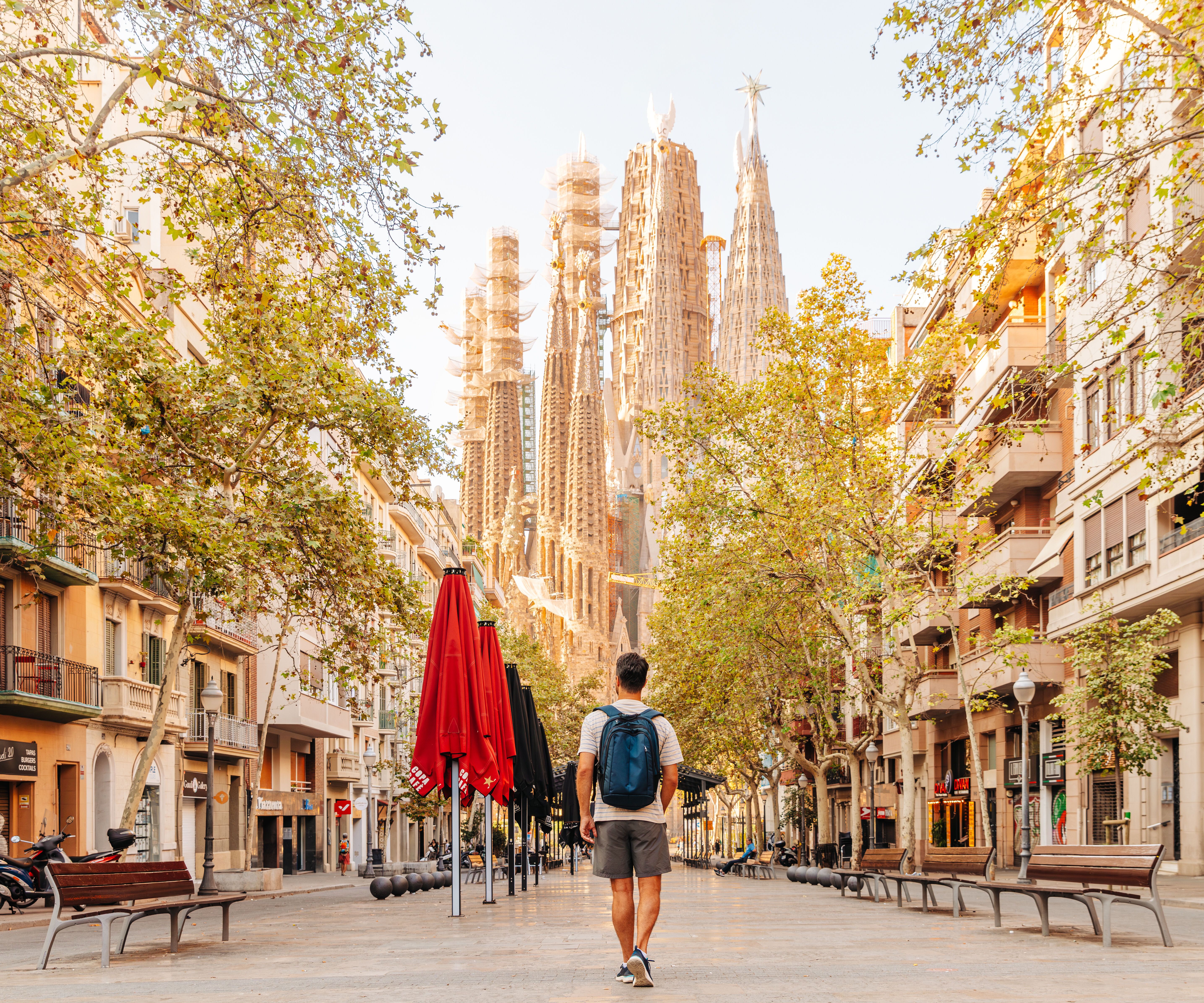 Ein Mann mit Rucksack geht an einem sonnigen Tag auf einer baumgesäumten Straße in Barcelona, mit Blick auf die markante Silhouette der Sagrada Familia im Hintergrund.