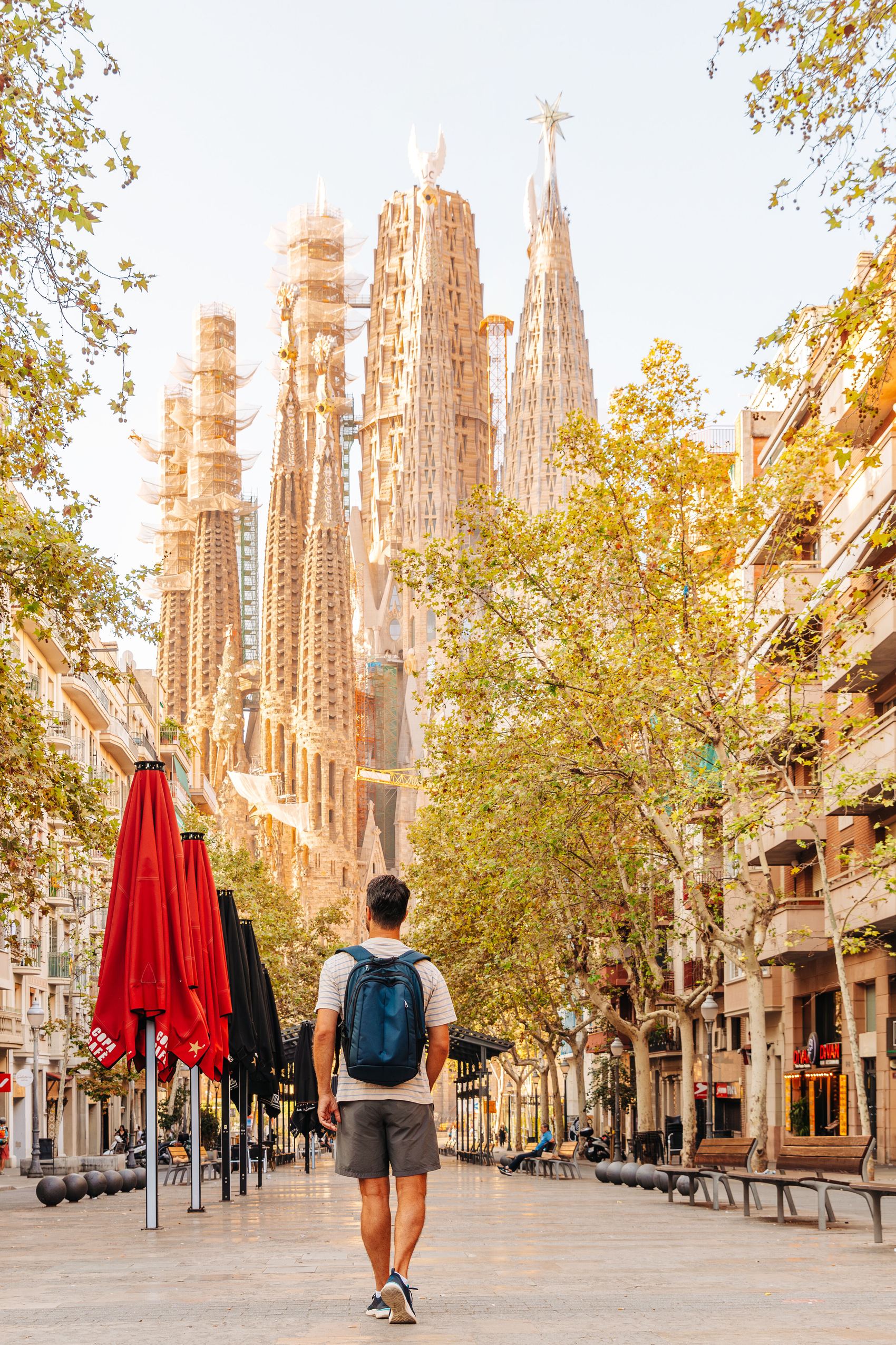 Ein Mann mit Rucksack geht an einem sonnigen Tag auf einer baumgesäumten Straße in Barcelona, mit Blick auf die markante Silhouette der Sagrada Familia im Hintergrund.