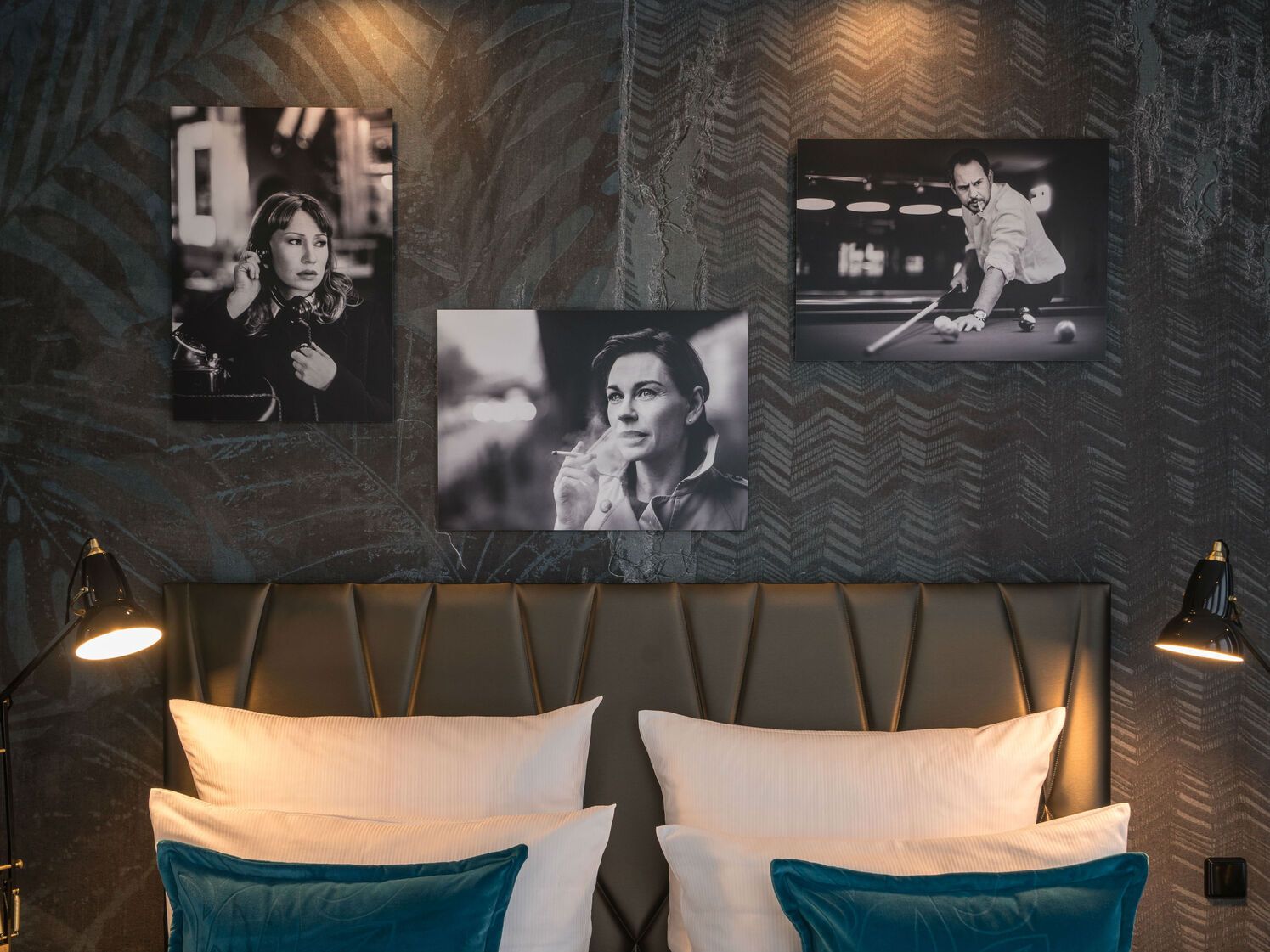 Elegantes Hotelzimmer mit gemütlichem Doppelbett, blauen Kissen und stilvoller Beleuchtung. An der Wand hängen drei Schwarz-Weiß-Fotografien, die eine nachdenkliche Frau und einen Mann beim Billard zeigen, was dem Raum eine künstlerische Atmosphäre verleiht.