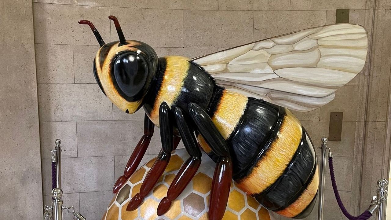 Große, realistisch gestaltete Bienenfigur mit schwarzen und gelben Streifen sitzt auf einer künstlerisch gestalteten Wabe, umgeben von Absperrungen in einem Innenraum.