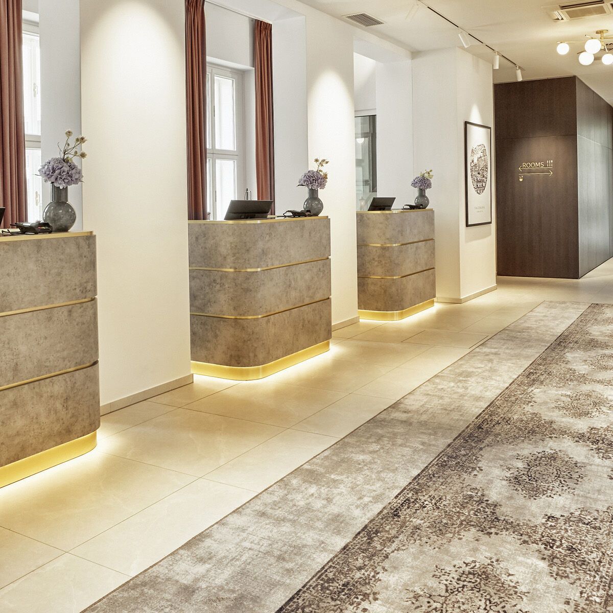 Heller, moderner Hotelempfang mit drei stilvollen Rezeptionstresen, warmem Licht, hohen Fenstern mit roten Vorhängen und elegantem Teppichboden, der eine einladende und professionelle Atmosphäre für Gäste schafft.