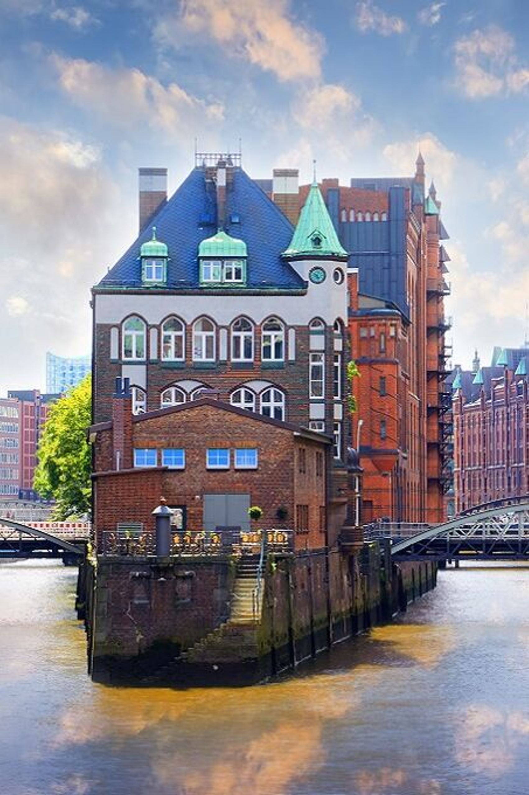 Blick auf das historische Wasserschloss in Hamburg nahe Hotels Hamburg, umgeben von roten Backsteingebäuden der Speicherstadt, mit zwei Brücken über einen ruhigen Kanal unter einem bewölkten Himmel.