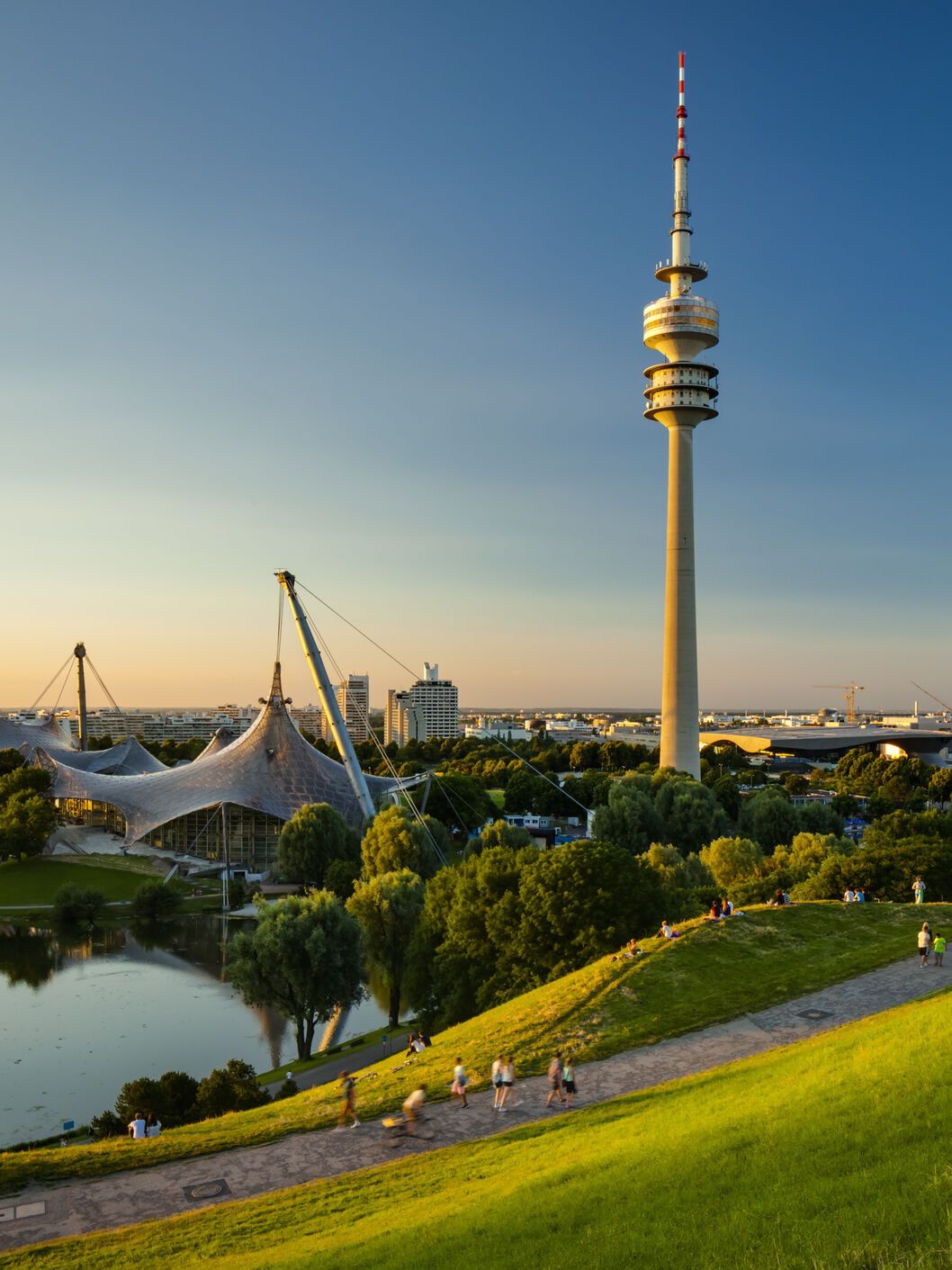 Blick über eine grüne Parklandschaft mit See und Spazierwegen, im Hintergrund der markante Olympiaturm und das Zeltdach des Olympiaparks bei klarem, sonnigem Himmel.