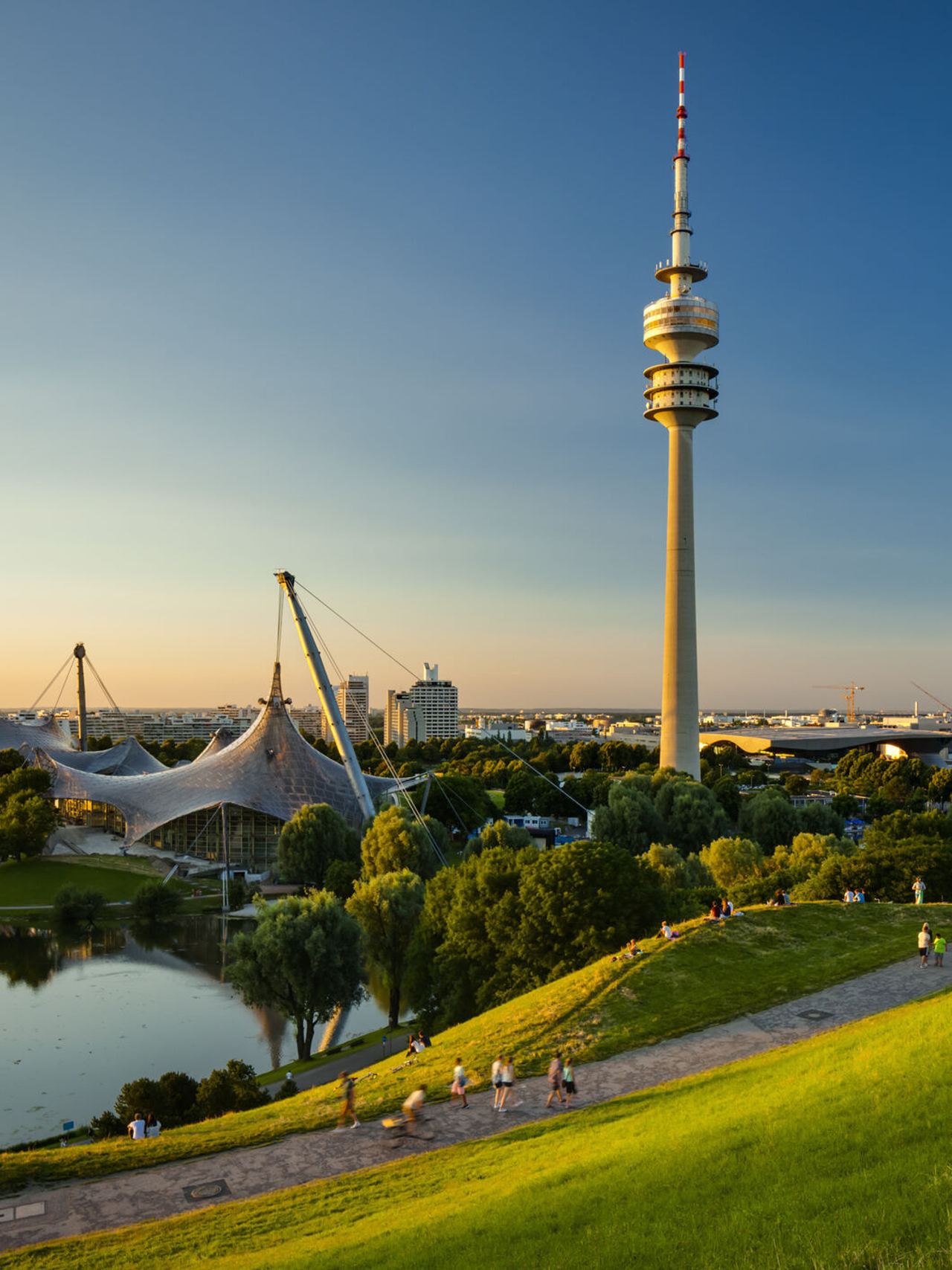 Blick über eine grüne Parklandschaft mit See und Spazierwegen, im Hintergrund der markante Olympiaturm und das Zeltdach des Olympiaparks bei klarem, sonnigem Himmel.