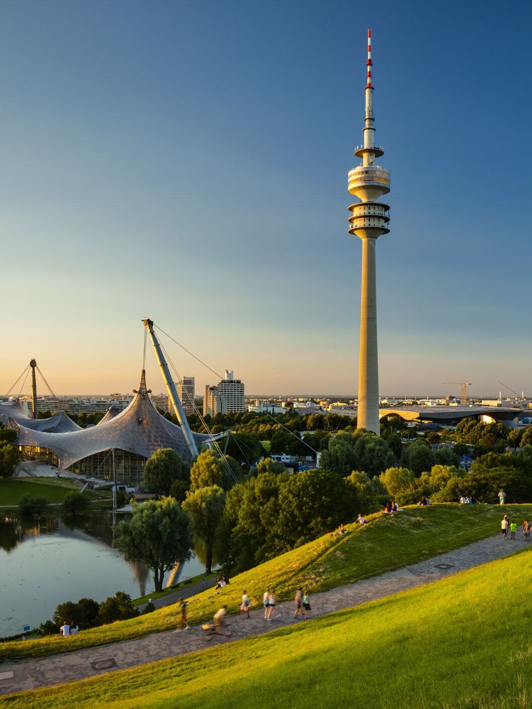 Blick über eine grüne Parklandschaft mit See und Spazierwegen, im Hintergrund der markante Olympiaturm und das Zeltdach des Olympiaparks bei klarem, sonnigem Himmel.