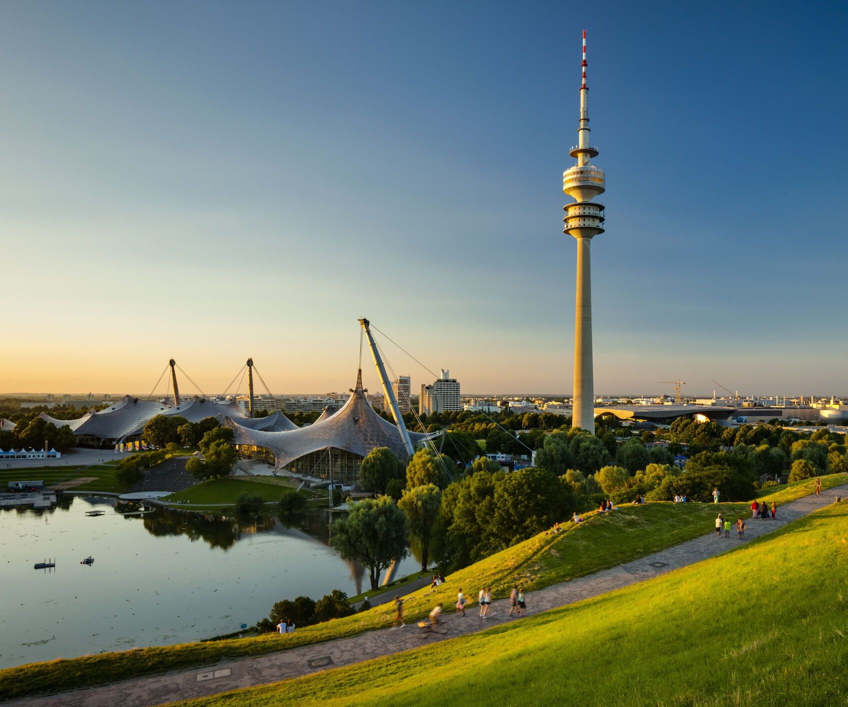 Blick über eine grüne Parklandschaft mit See und Spazierwegen, im Hintergrund der markante Olympiaturm und das Zeltdach des Olympiaparks bei klarem, sonnigem Himmel.