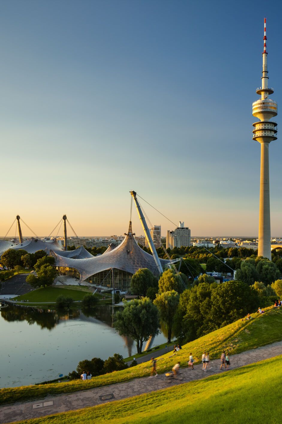 Blick über eine grüne Parklandschaft mit See und Spazierwegen, im Hintergrund der markante Olympiaturm und das Zeltdach des Olympiaparks bei klarem, sonnigem Himmel.