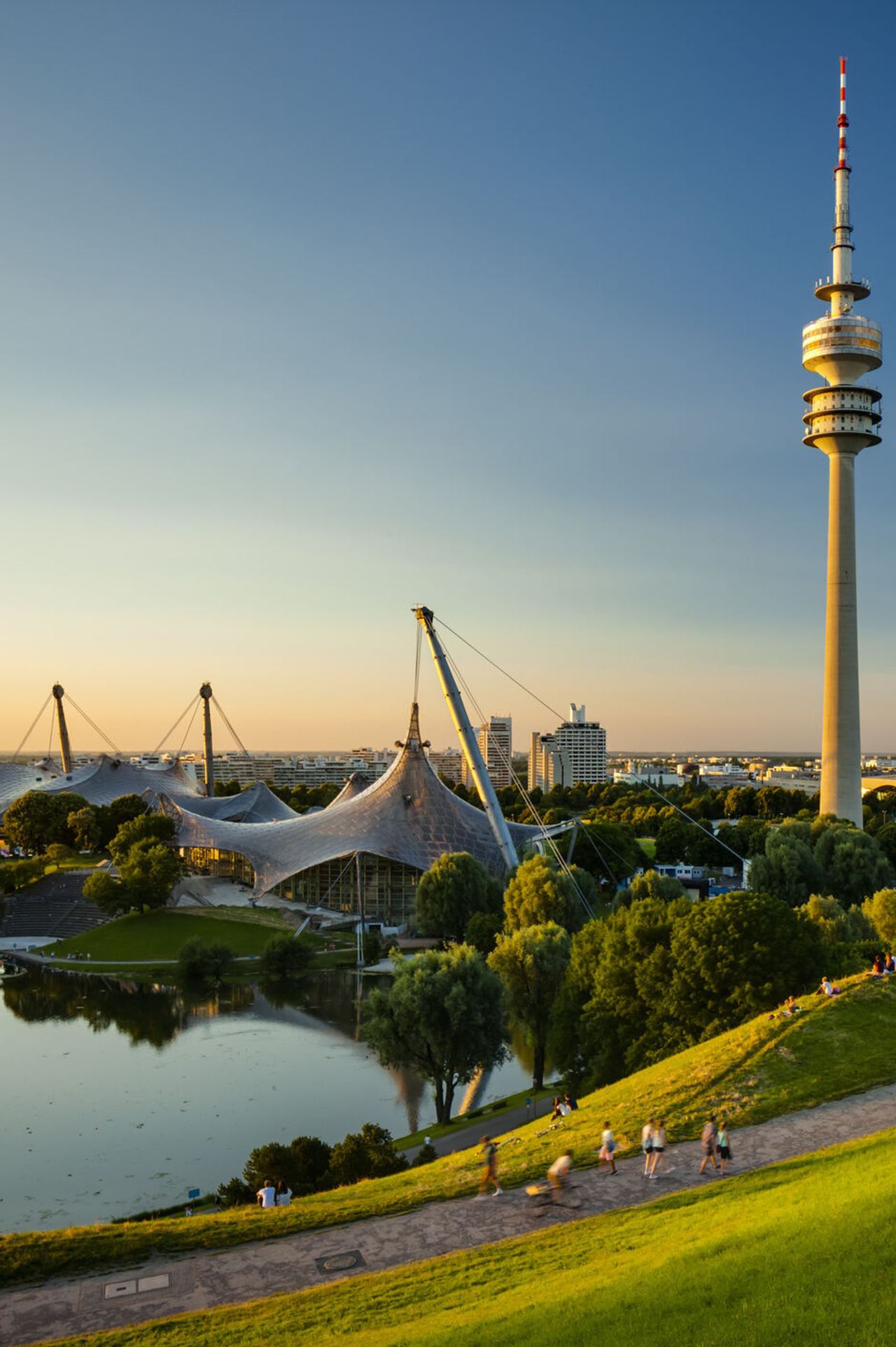 Blick über eine grüne Parklandschaft mit See und Spazierwegen, im Hintergrund der markante Olympiaturm und das Zeltdach des Olympiaparks bei klarem, sonnigem Himmel. Blick über eine grüne Parklandschaft mit See und Spazierwegen, im Hintergrund der markante Olympiaturm und das Zeltdach des Olympiaparks bei klarem, sonnigem Himmel.