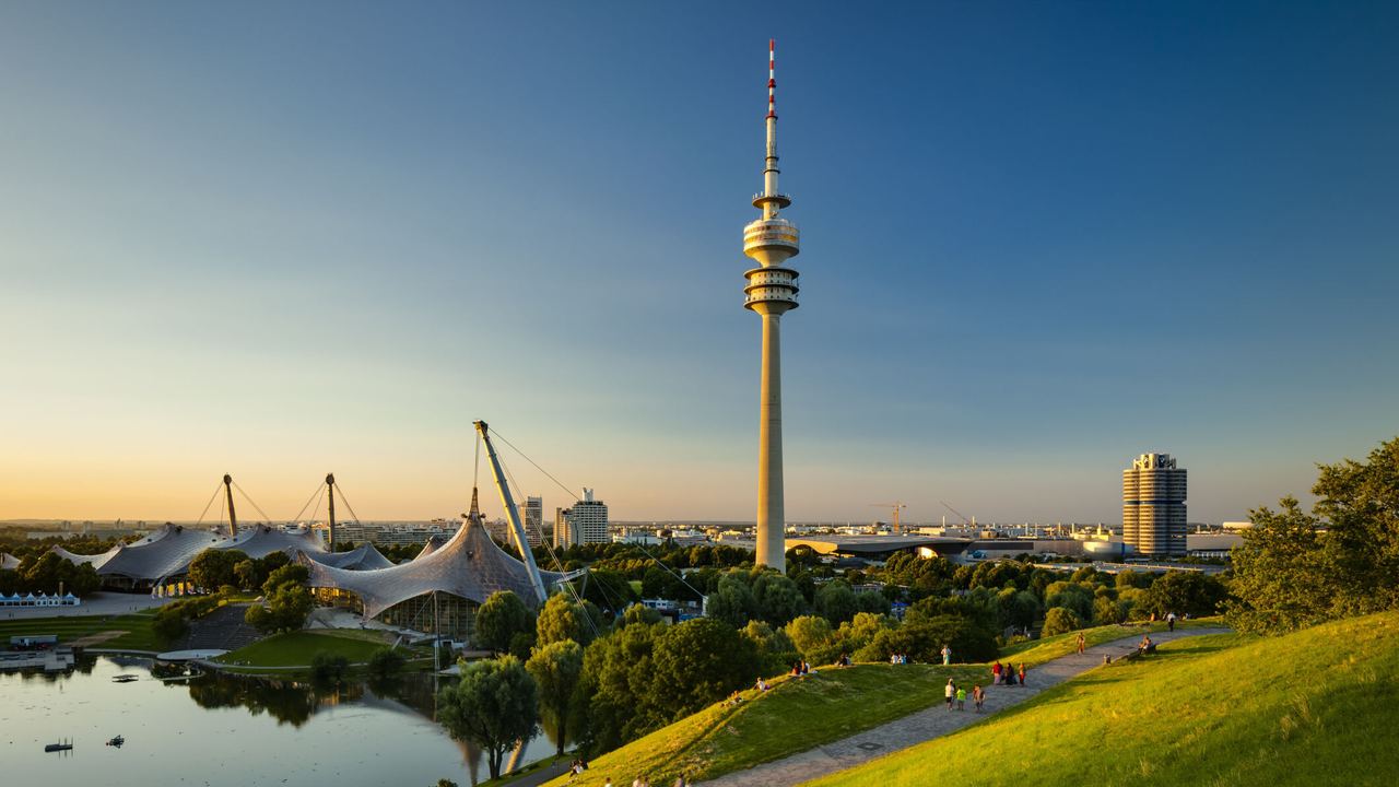 Blick über eine grüne Parklandschaft mit See und Spazierwegen, im Hintergrund der markante Olympiaturm und das Zeltdach des Olympiaparks bei klarem, sonnigem Himmel.