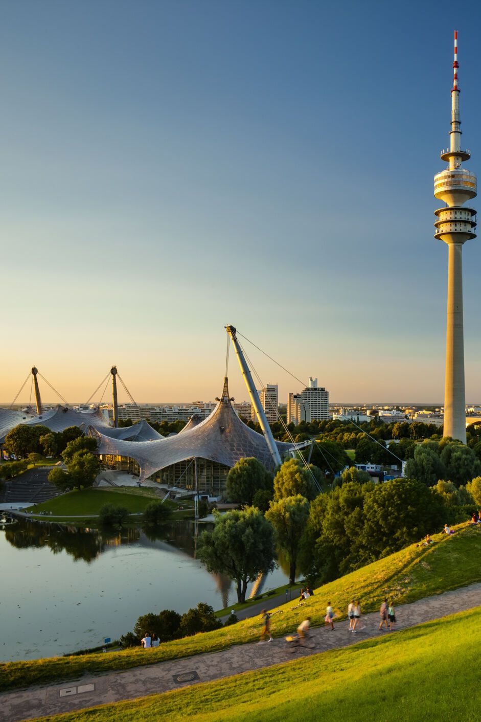 Blick über eine grüne Parklandschaft mit See und Spazierwegen, im Hintergrund der markante Olympiaturm und das Zeltdach des Olympiaparks bei klarem, sonnigem Himmel.