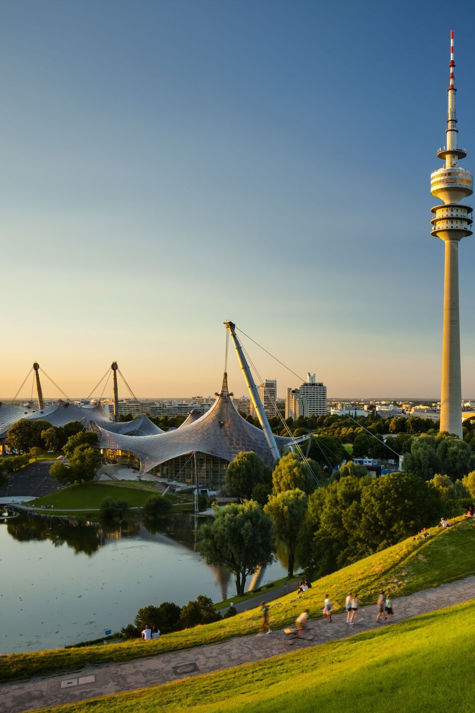 Blick über eine grüne Parklandschaft mit See und Spazierwegen, im Hintergrund der markante Olympiaturm und das Zeltdach des Olympiaparks bei klarem, sonnigem Himmel.