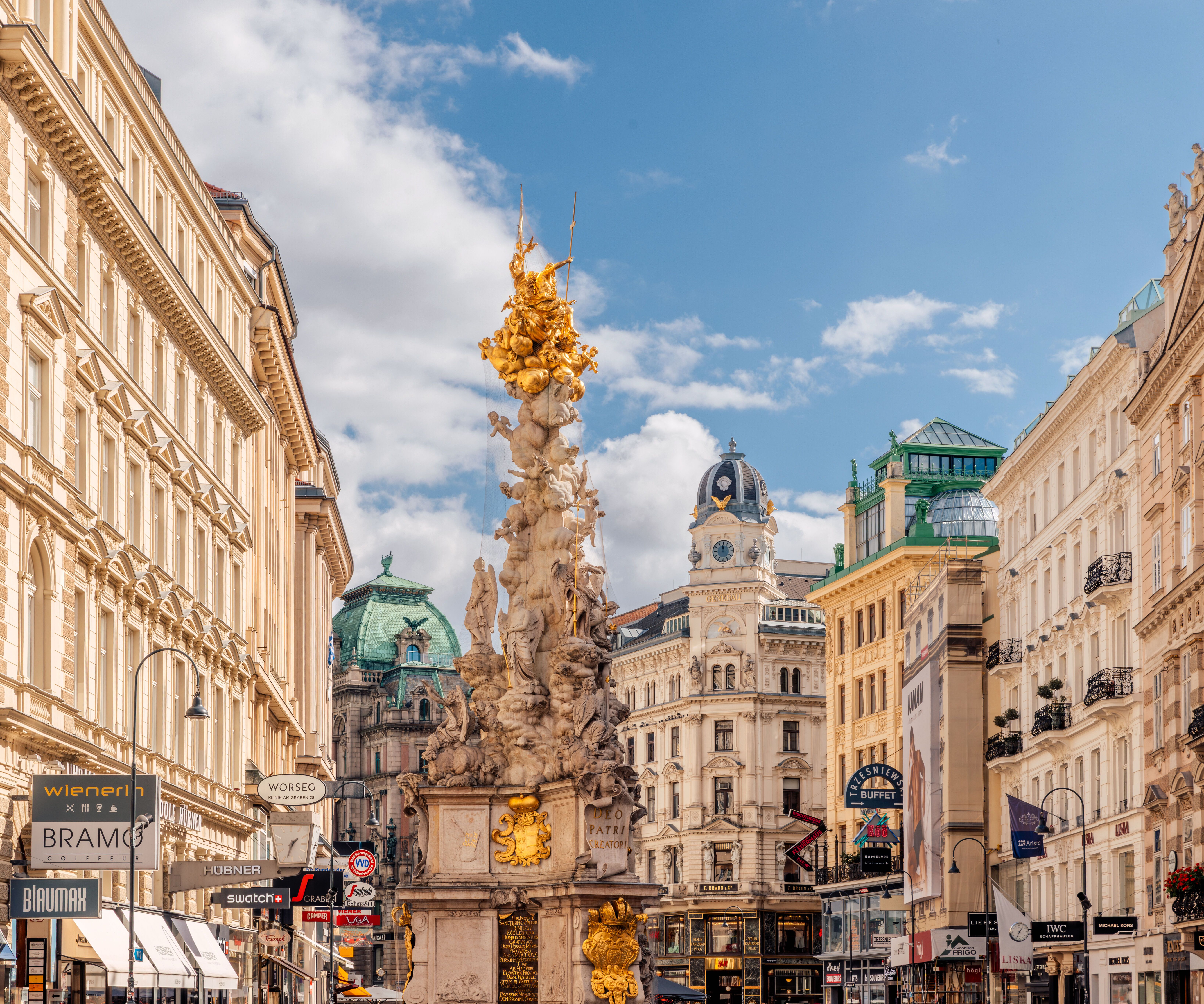 Historische Wiener Innenstadt mit prächtigen barocken Gebäuden, zentraler kunstvoll verzierter Säule und lebhaftem Straßenbild unter blauem Himmel.