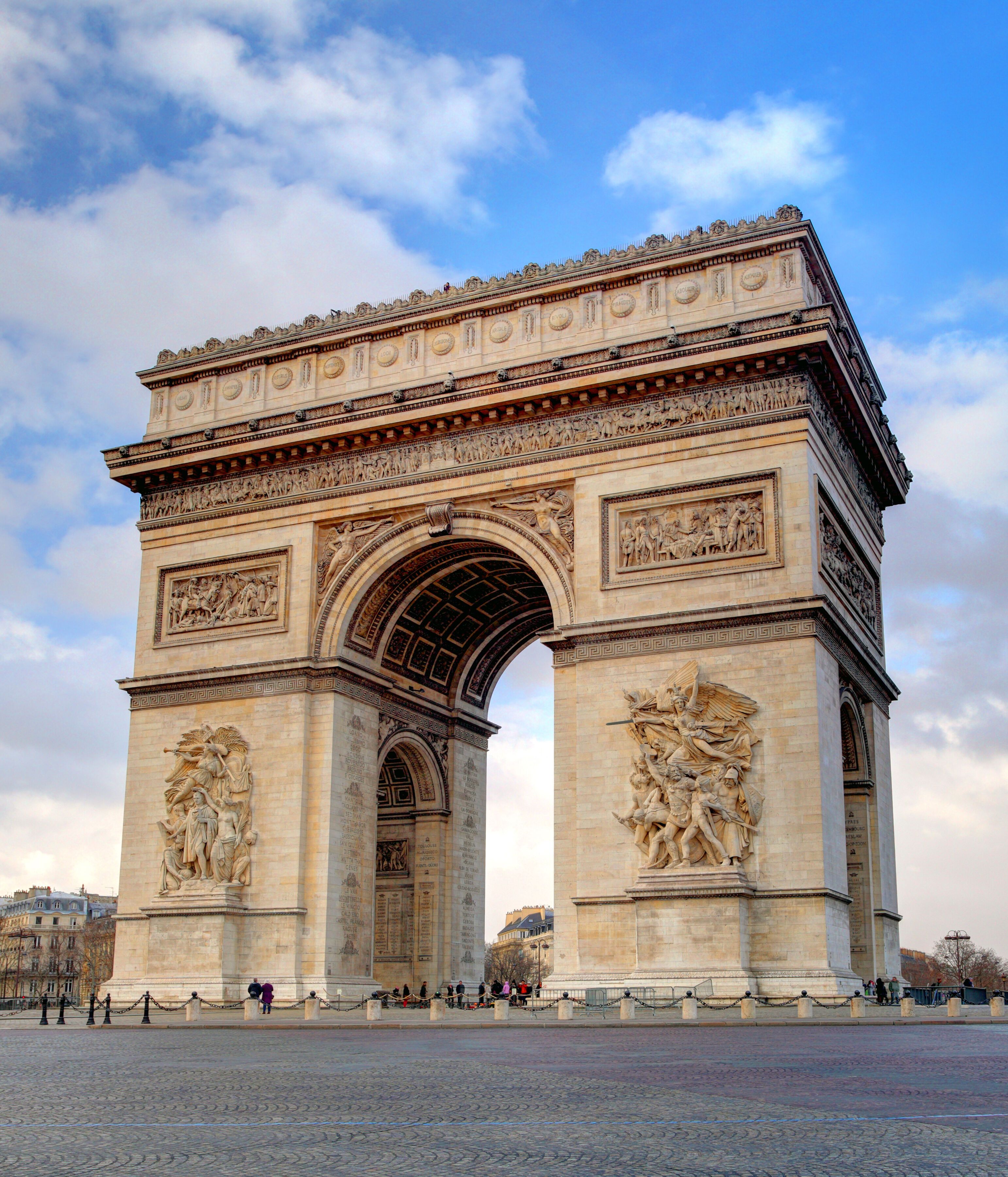 Der Arc de Triomphe in Paris, ein prächtiges, historisches Monument mit Reliefs, steht auf einem großen, offenen Platz unter blauem Himmel mit Wolken.