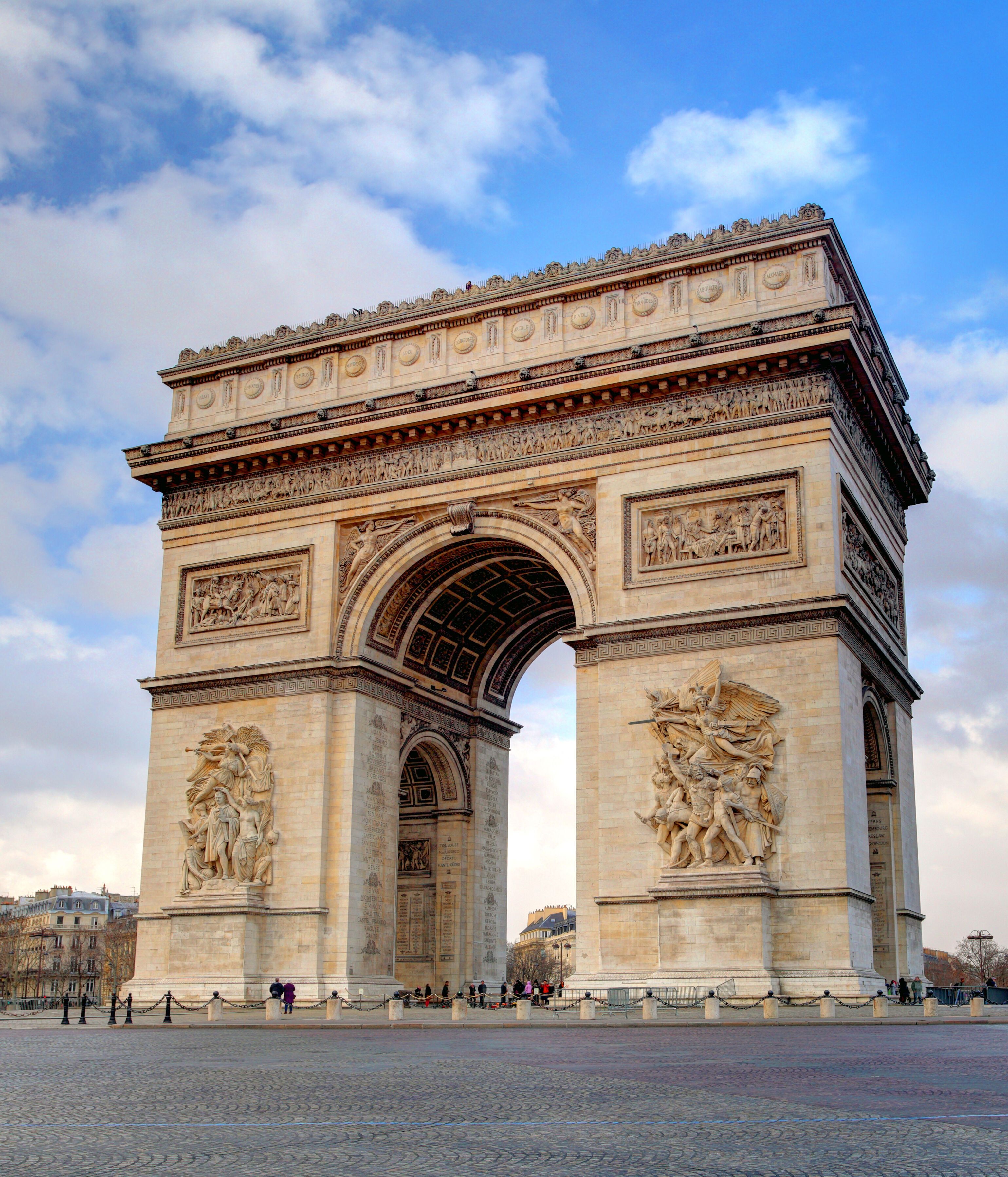 Der Arc de Triomphe in Paris, ein prächtiges, historisches Monument mit Reliefs, steht auf einem großen, offenen Platz unter blauem Himmel mit Wolken.