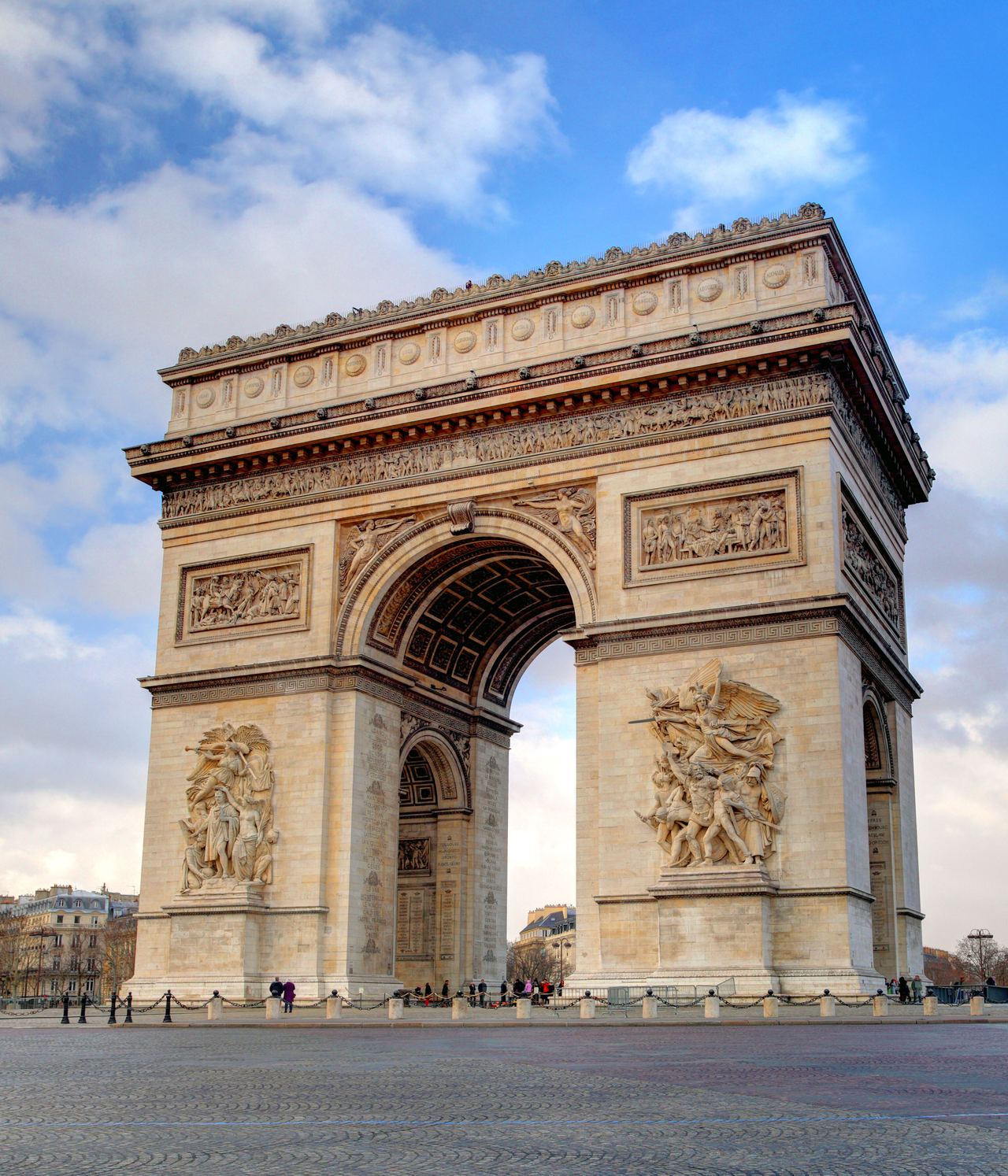 Der Arc de Triomphe in Paris, ein prächtiges, historisches Monument mit Reliefs, steht auf einem großen, offenen Platz unter blauem Himmel mit Wolken.
