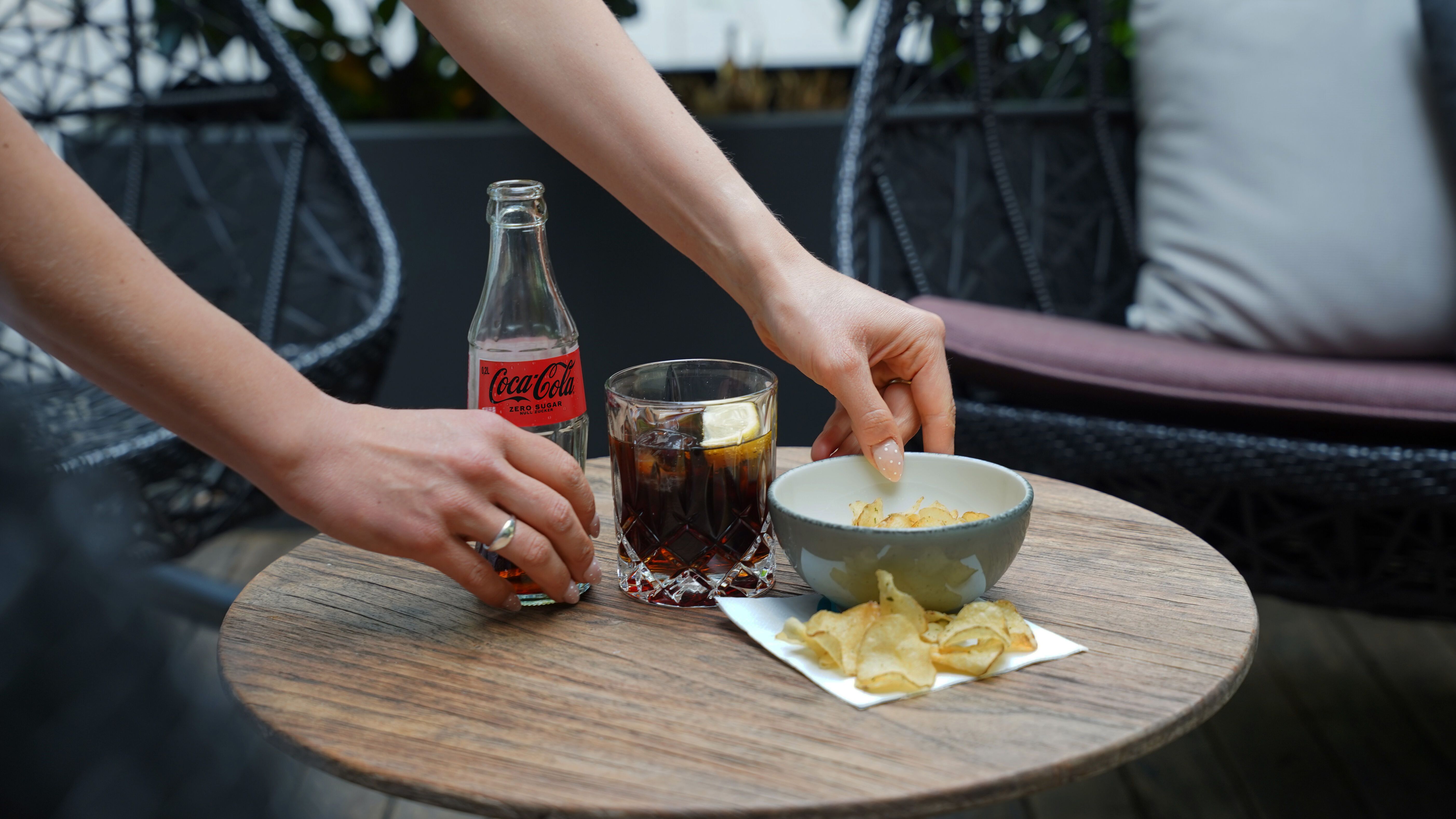 Eine Person greift nach Kartoffelchips aus einer Schale auf einem runden Holztisch, daneben steht ein Glas Cola mit Zitronenscheibe und eine offene Coca-Cola-Flasche.
