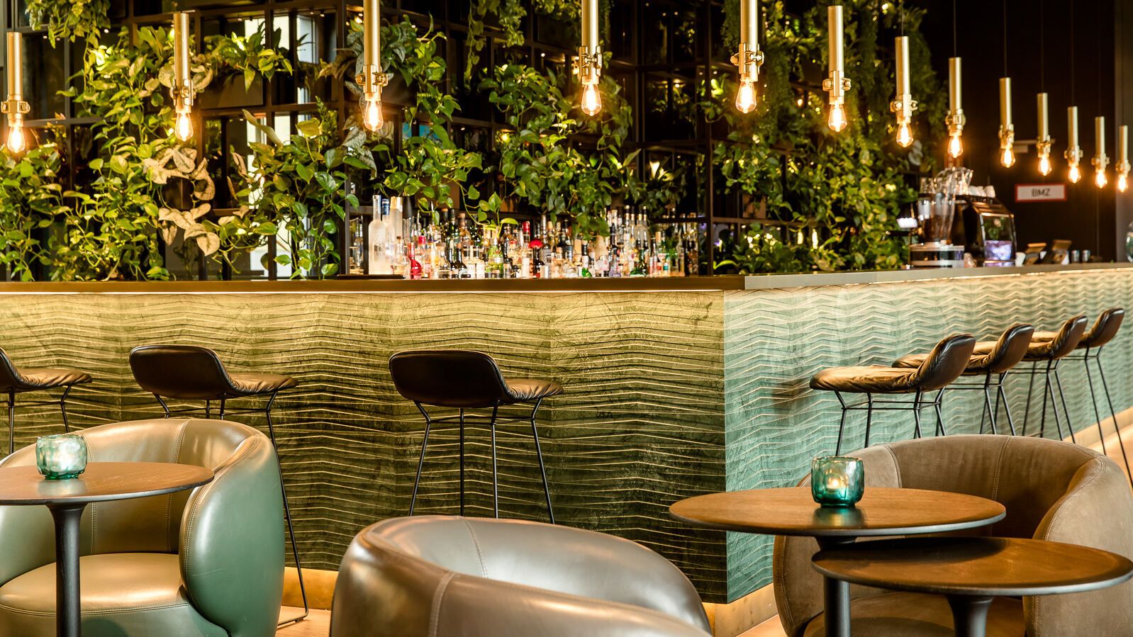 Motel One Hotelbar mit stilvoller grüner Wandbegrünung, warmem Licht von hängenden Glühbirnen und Sitzgelegenheiten aus Leder.