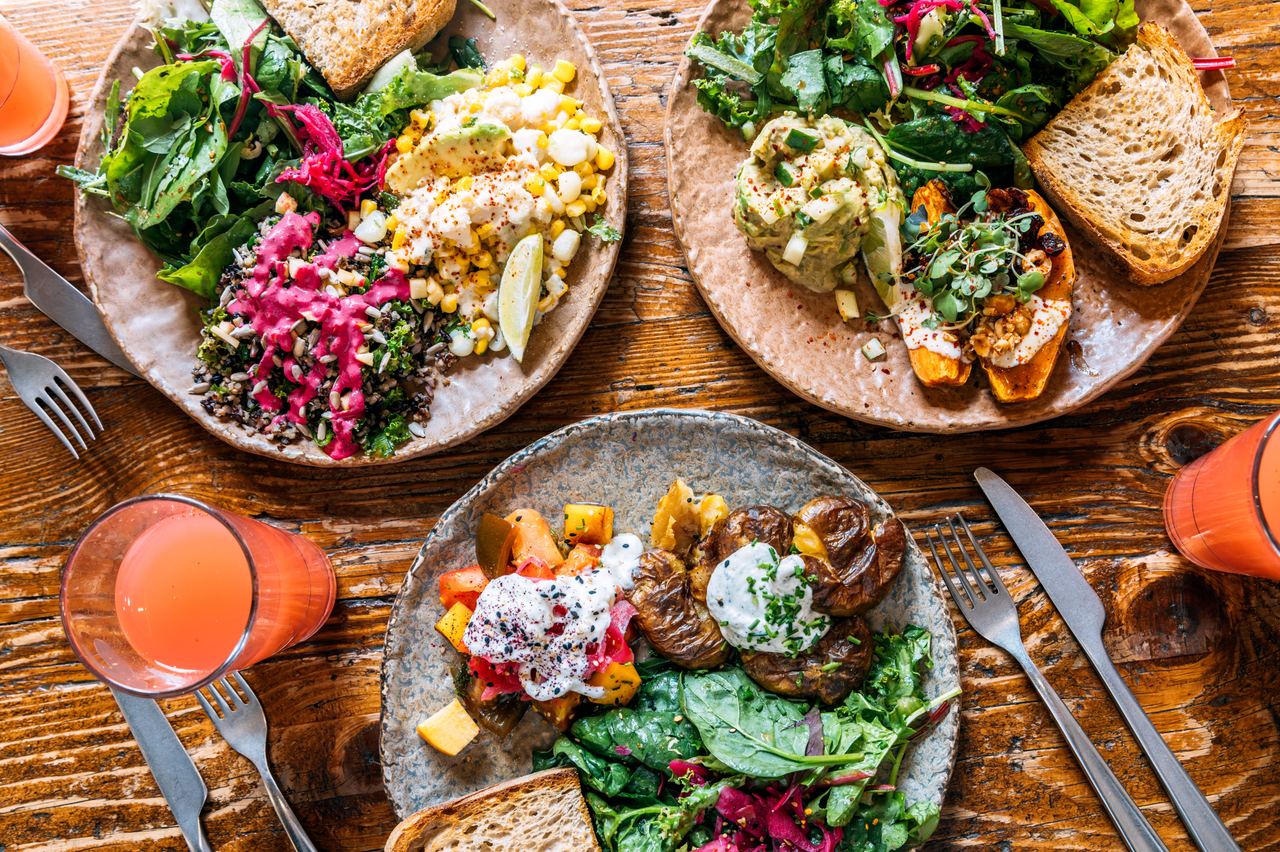 Drei Teller mit abwechslungsreichen, frischen Salaten, Gemüse, Kräutern und Vollkornbrot auf einem Holztisch, begleitet von Gläsern mit frisch gepresstem Orangensaft.