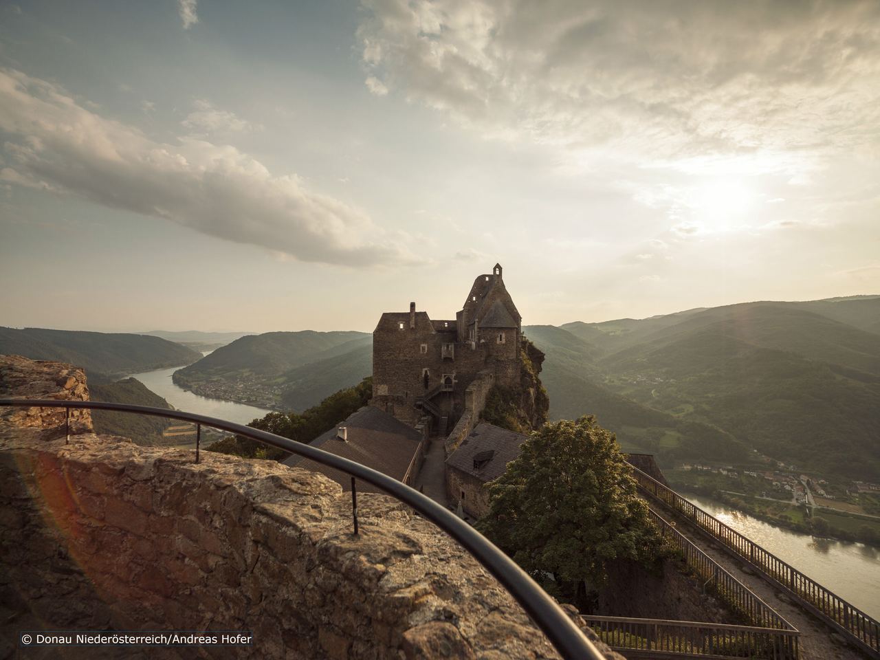 Blick auf eine historische Burg auf einem Hügel mit Blick ins Tal und einen Fluss, umgeben von bewaldeten Bergen bei Sonnenuntergang.